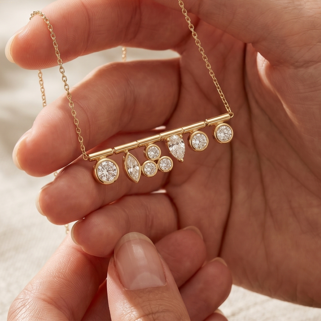 A gold bar necklace with multiple clear round and teardrop-cut gemstones, held gently in a person's hand against a soft light background.