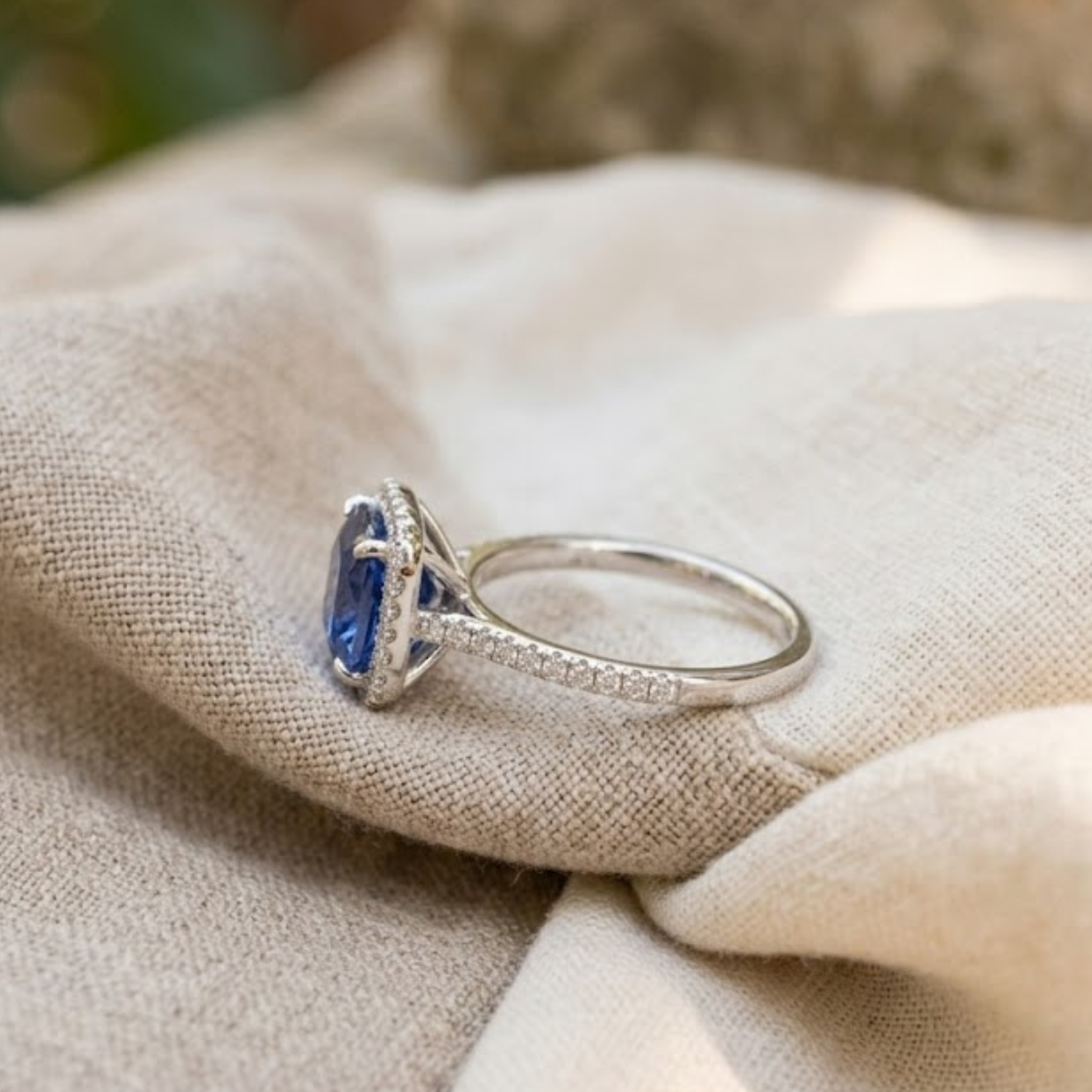 A silver ring with a blue oval gemstone and small clear stones along the band, resting on draped beige fabric.