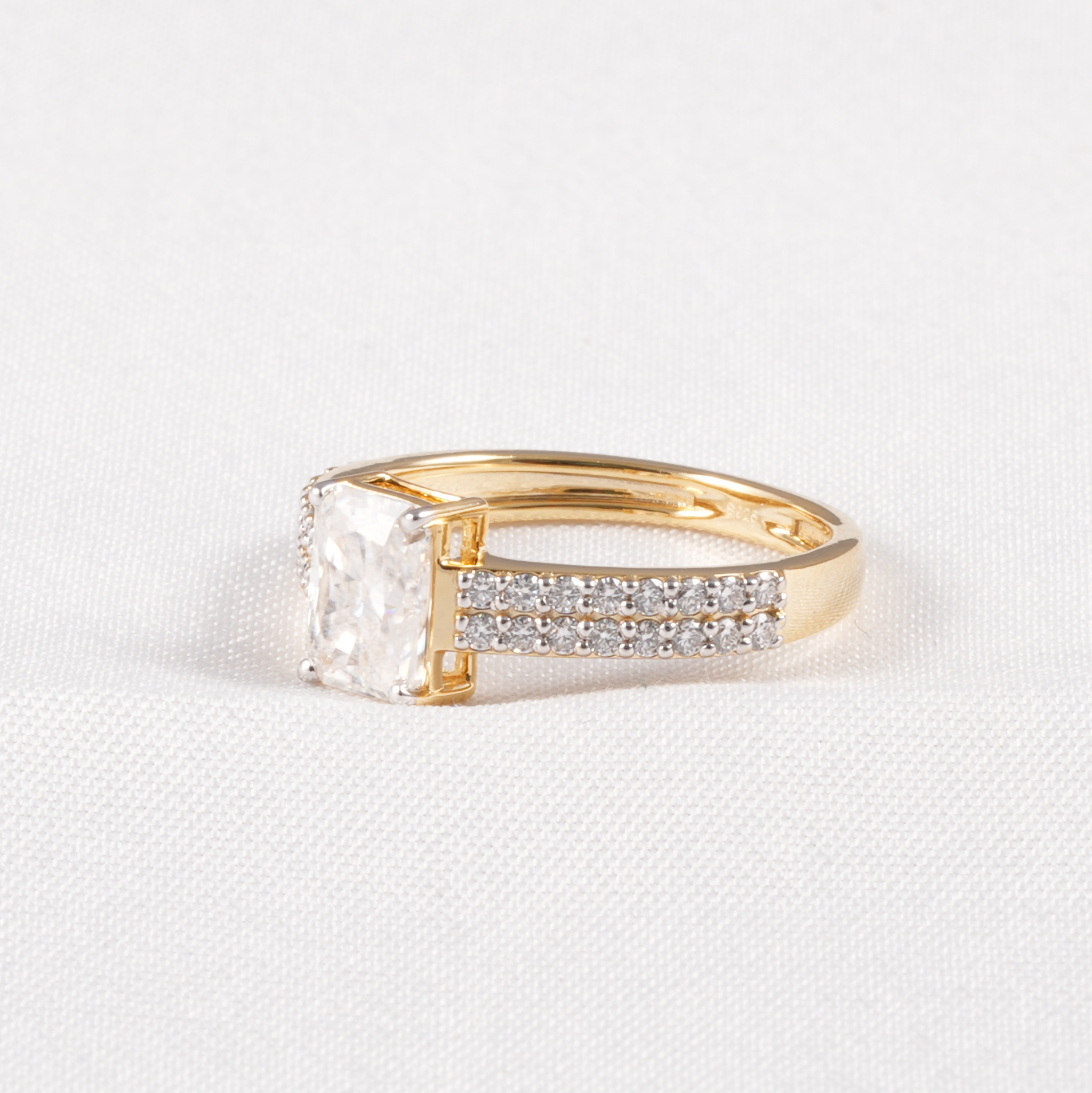 A yellow gold ring featuring a large rectangular clear gemstone and a pavé band with small clear stones, set against a white textured fabric background.