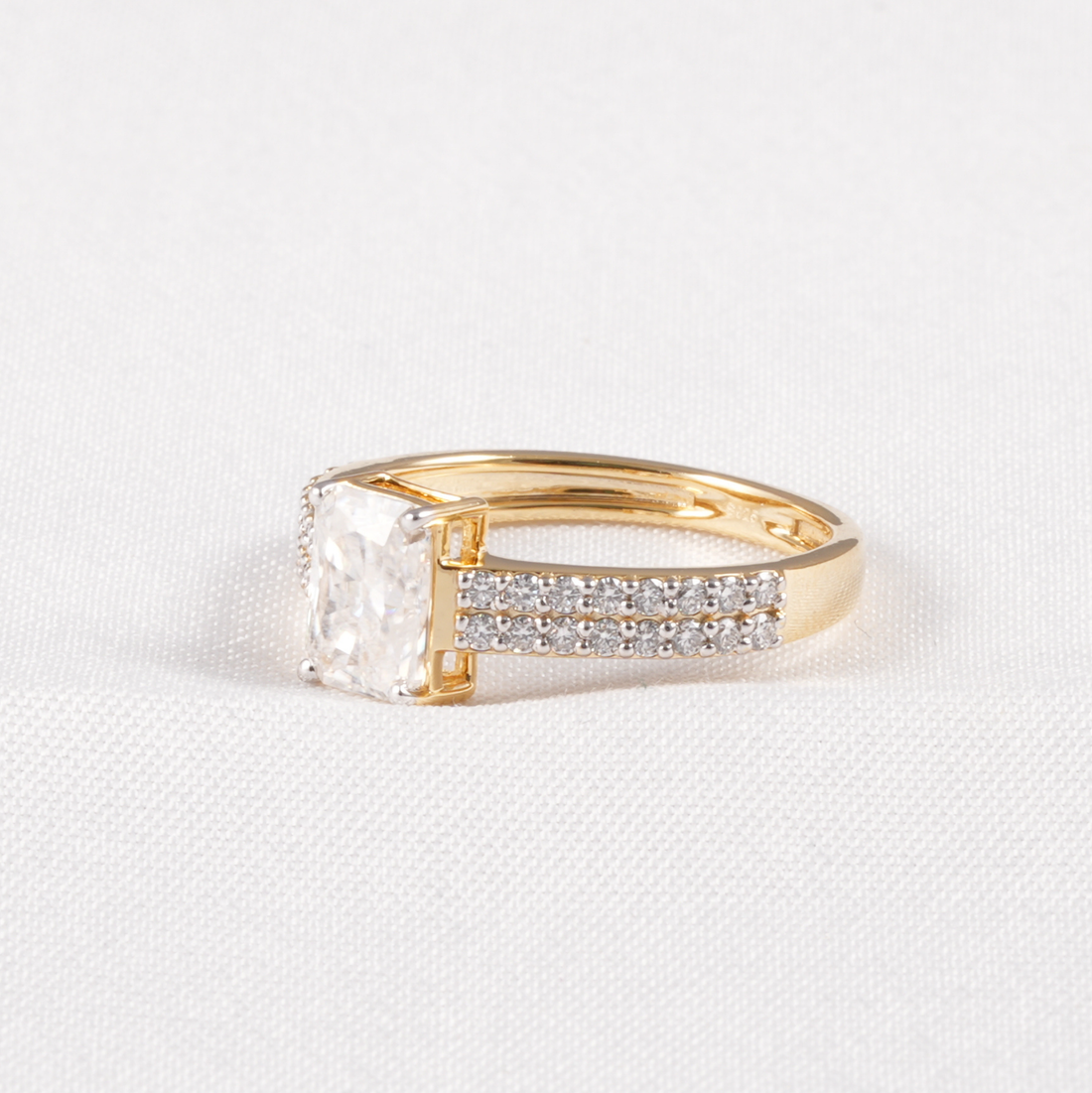 A yellow gold ring featuring a large rectangular clear gemstone and a pavé band with small clear stones, set against a white textured fabric background.