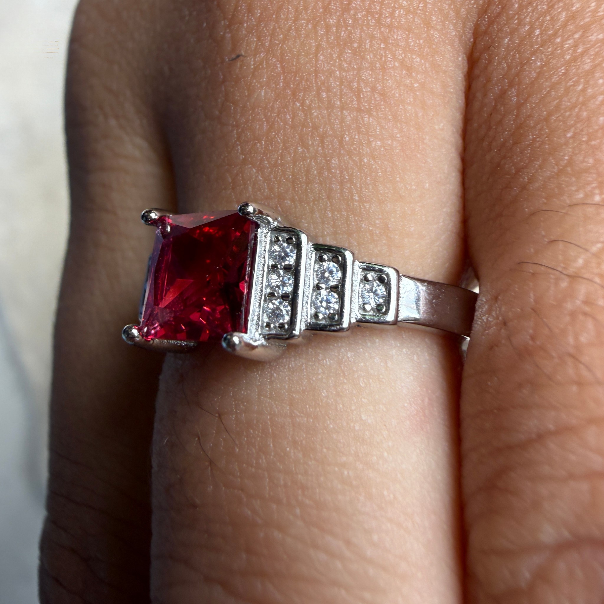 A silver ring with a large square-cut red gemstone and small clear accent stones, shown worn on a finger.