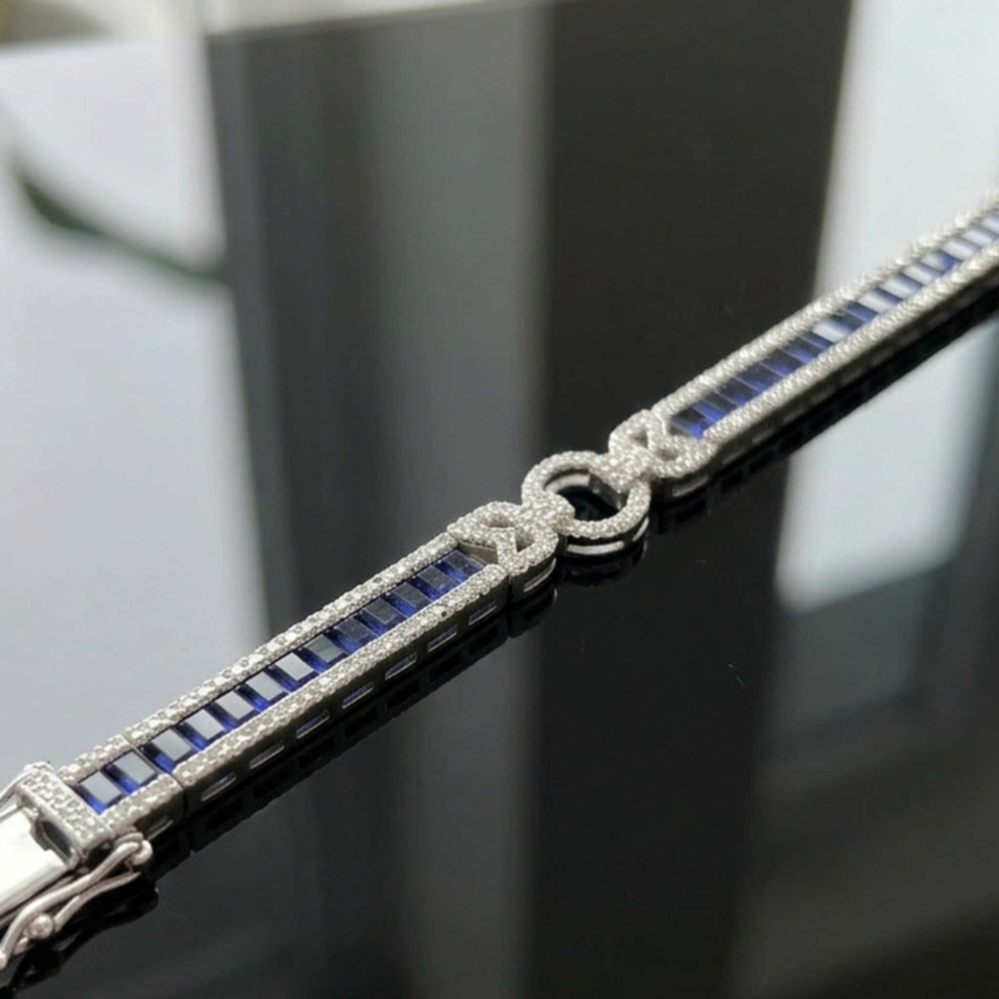 A close-up of a silver-toned bracelet featuring rectangular blue sapphires and pavé-set clear stones, including a central knot motif, displayed on a reflective dark surface.