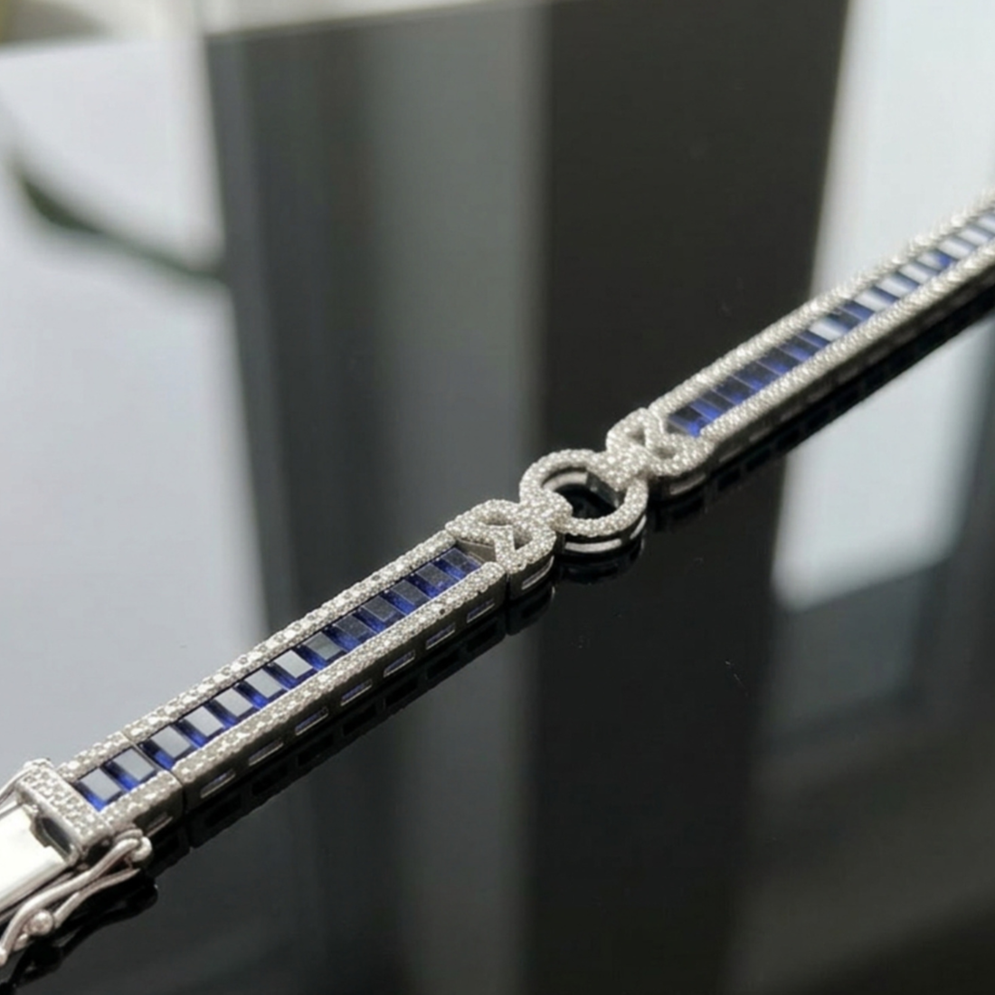 A close-up of a silver-toned bracelet featuring rectangular blue sapphires and pavé-set clear stones, including a central knot motif, displayed on a reflective dark surface.