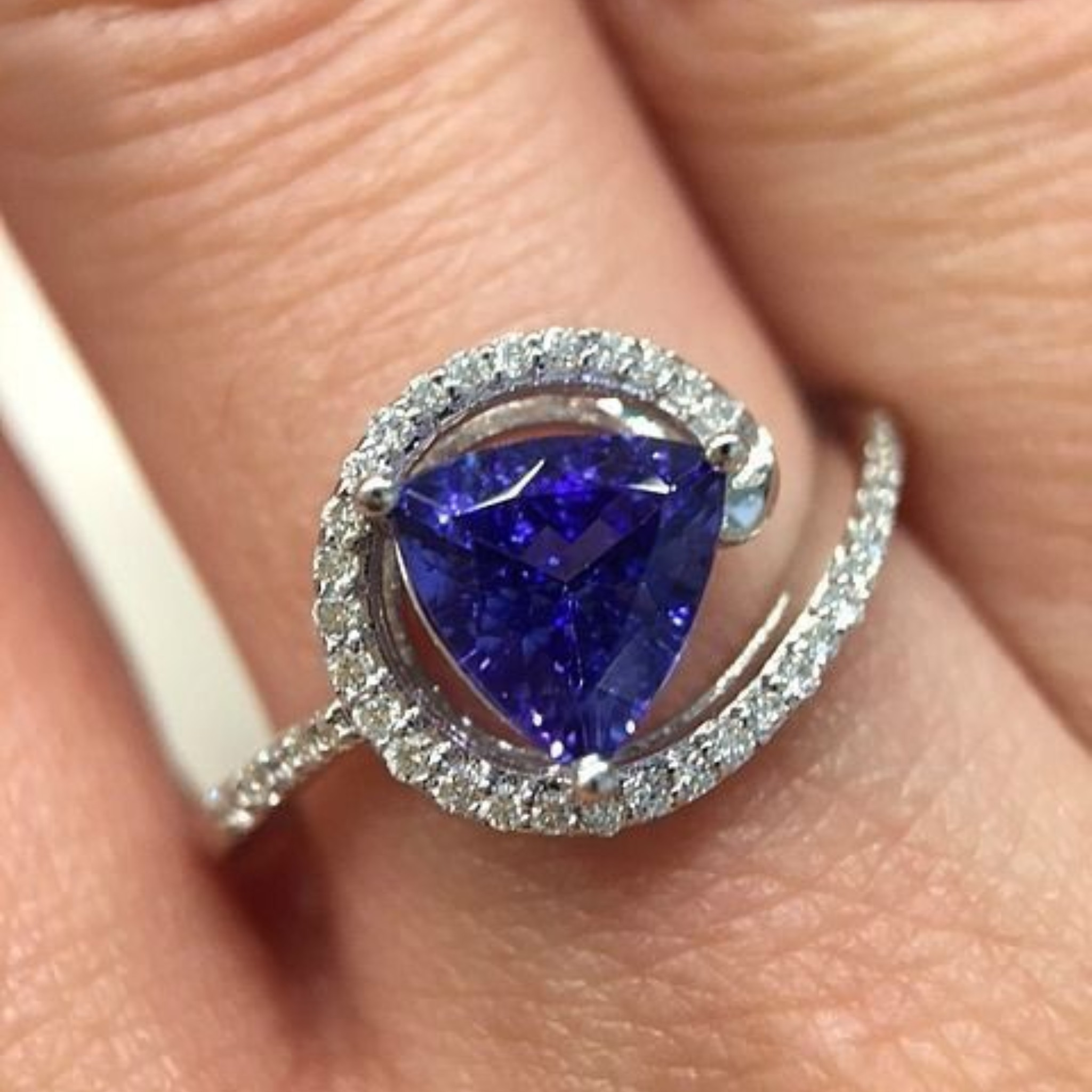 A close-up of a ring worn on a finger, featuring a large triangular deep blue gemstone surrounded by a halo of small clear stones and a curved silver-toned band with pavé-set accents.