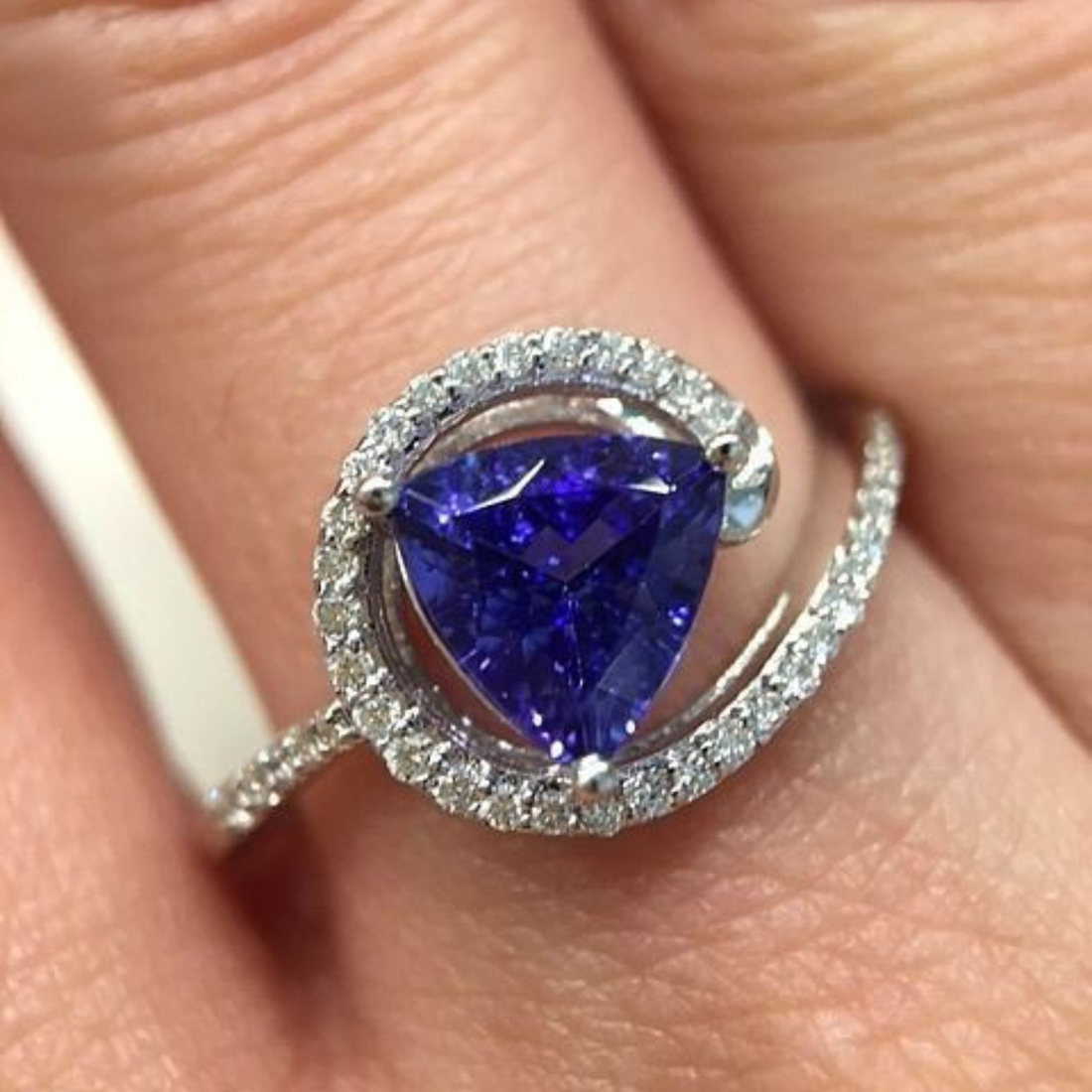 A close-up of a ring worn on a finger, featuring a large triangular deep blue gemstone surrounded by a halo of small clear stones and a curved silver-toned band with pavé-set accents.