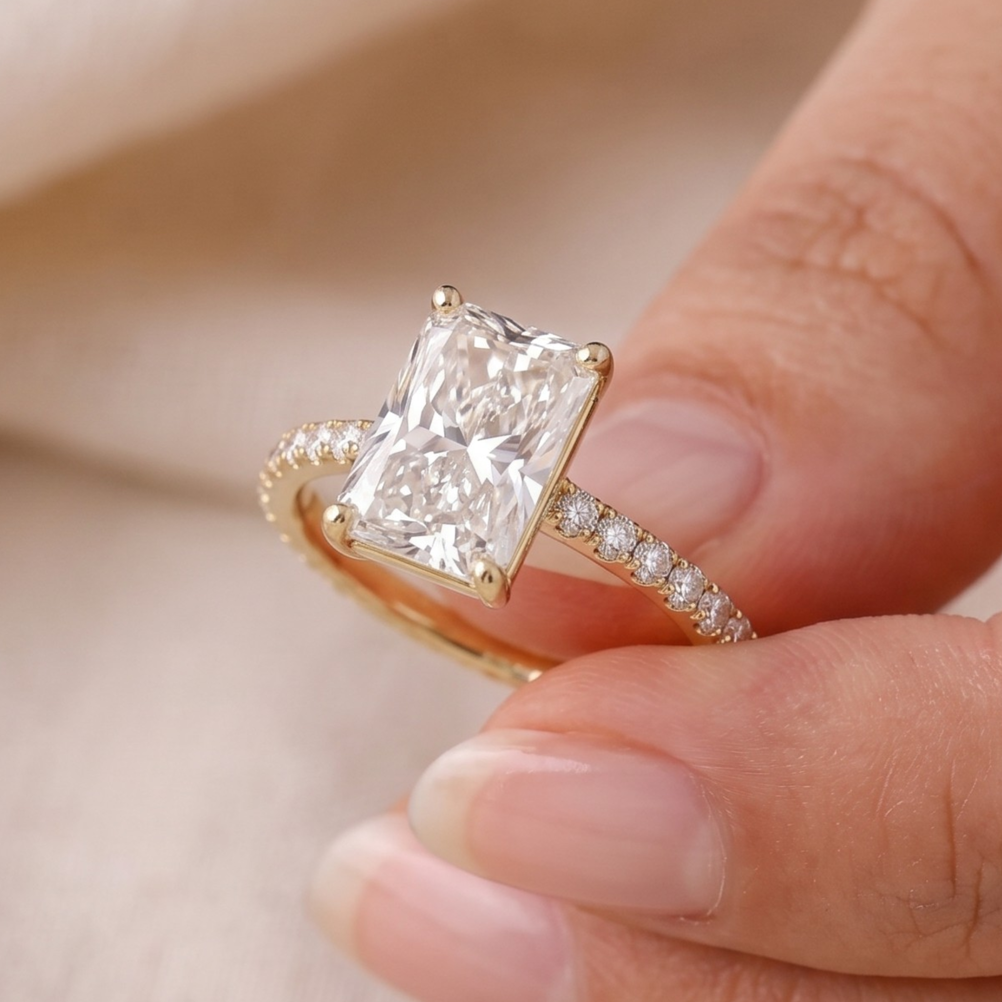 A close-up of a gold ring with a large rectangular clear gemstone and small side stones, shown being held between fingers against a soft beige background.