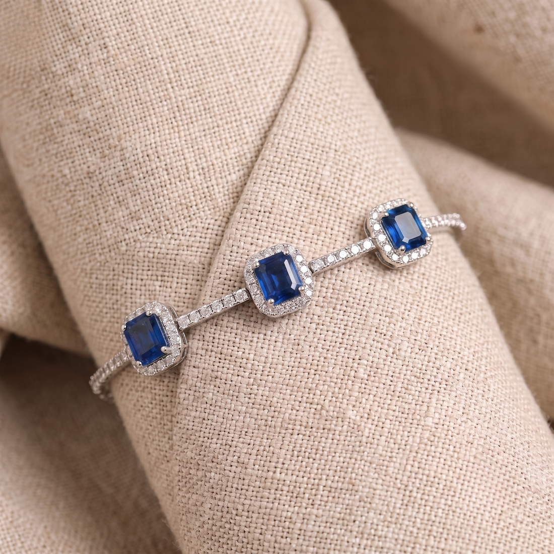 A silver-toned bracelet with three square-cut blue gemstones, each surrounded by a halo of small clear stones, displayed on a beige textured fabric surface.