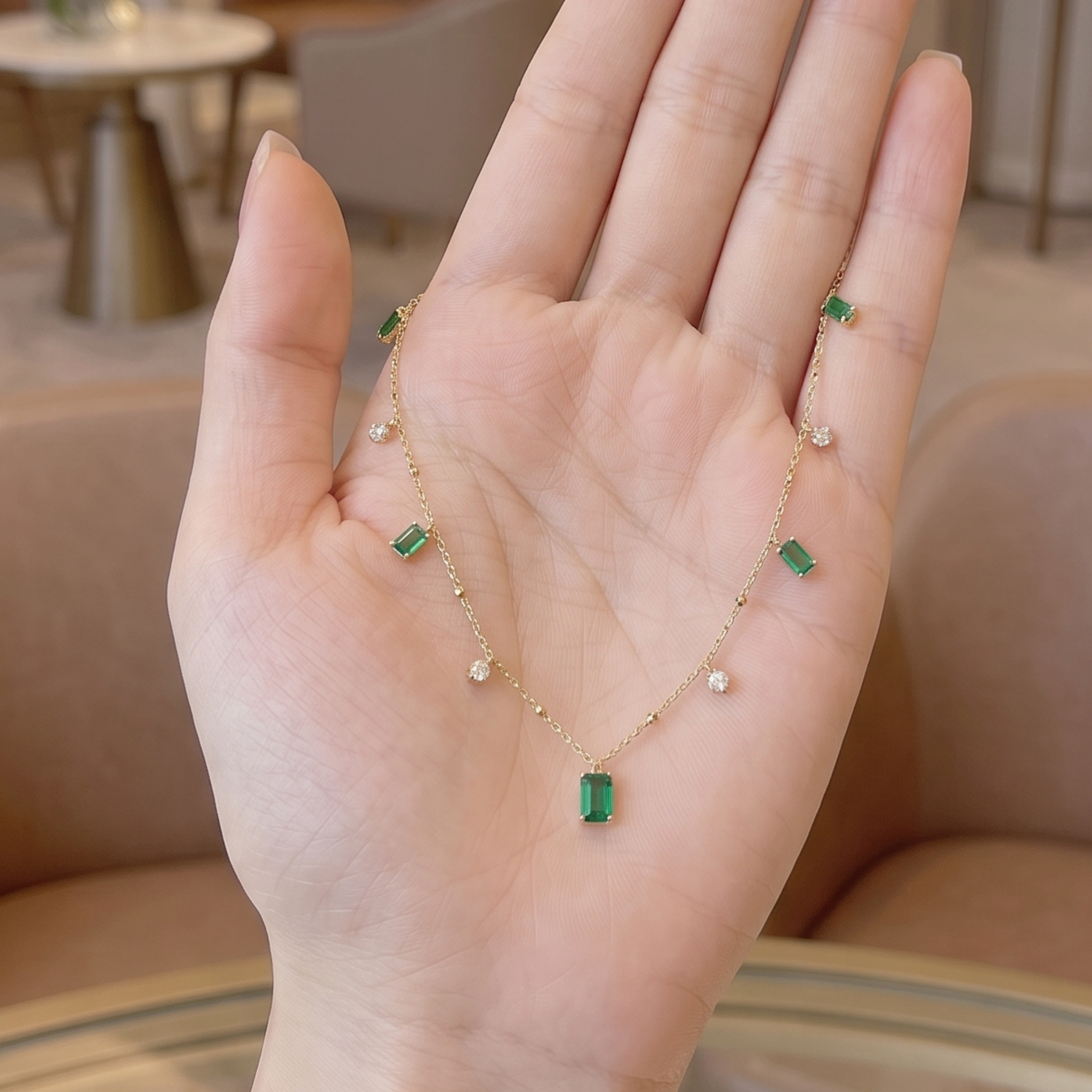 A delicate gold chain necklace with green rectangular and round gemstones, displayed on an open hand. The necklace features a central green rectangular pendant, smaller green rectangular accents, and small clear round stones spaced along the chain.