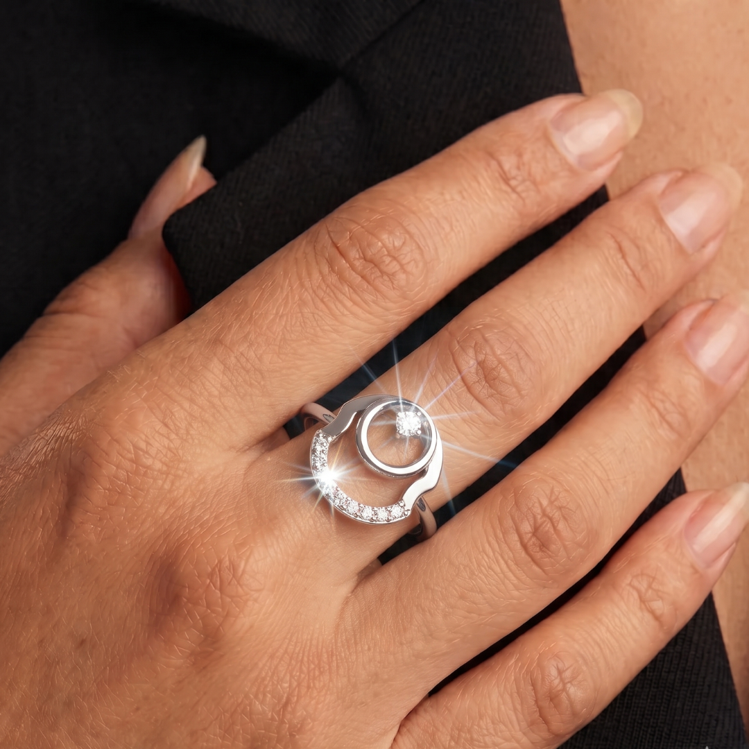 A close-up of a hand wearing a silver ring with a circular design and small clear stones, set against a dark sleeve and skin background.