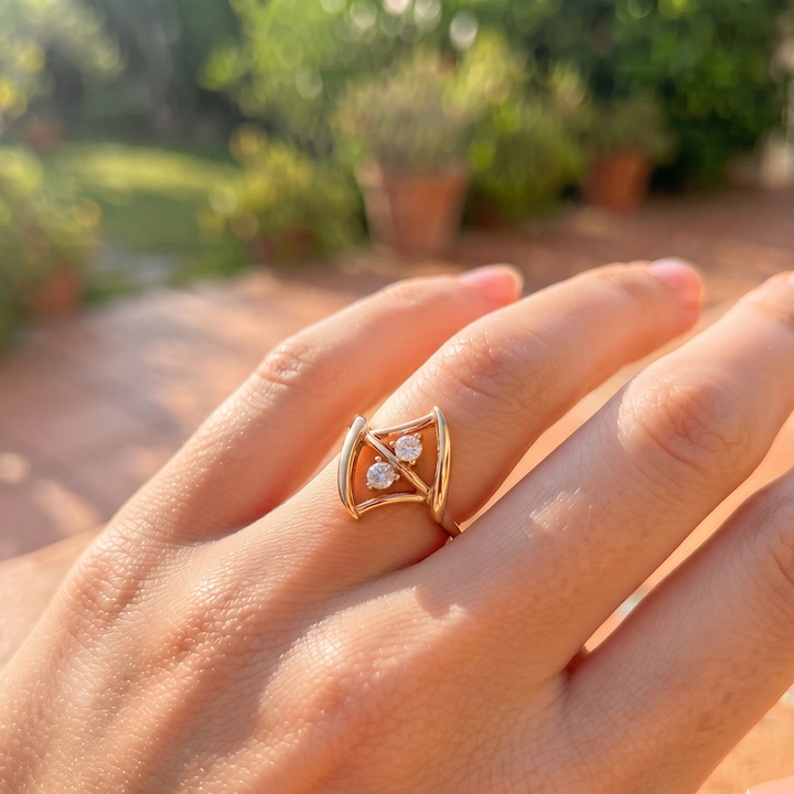 A gold ring with three small clear stones set in a triangular design, worn on a person's finger against a blurred outdoor garden background.
