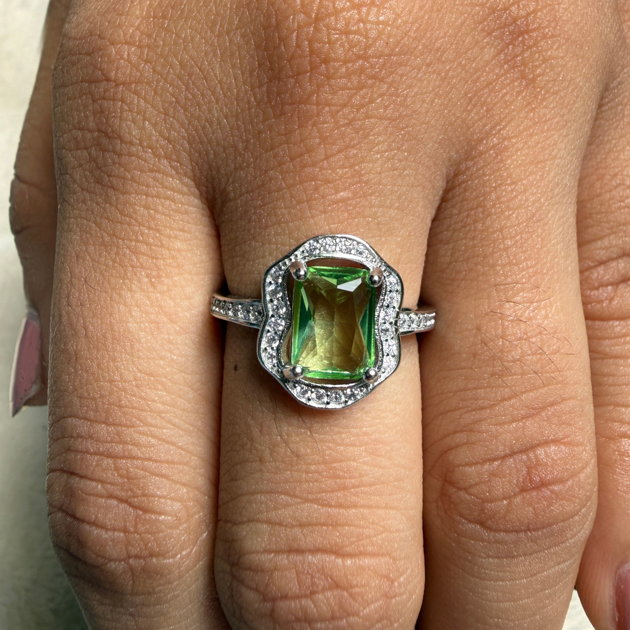 A silver ring with a green rectangular emerald-cut center stone and a halo of small clear stones, shown worn on a finger.