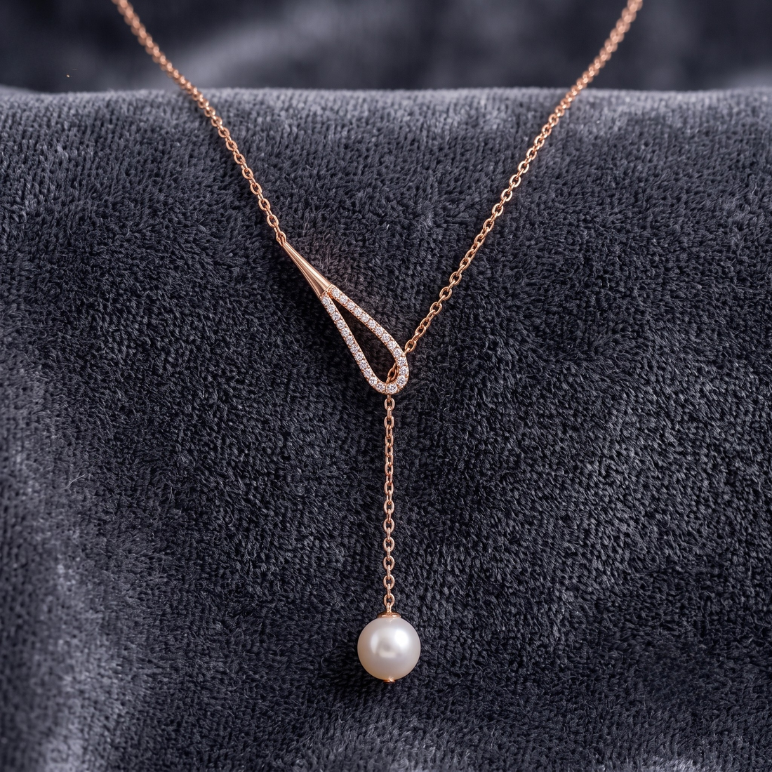 A rose gold Y-shaped necklace with a small diamond-accented loop and a dangling pearl, displayed on a dark textured velvet surface.