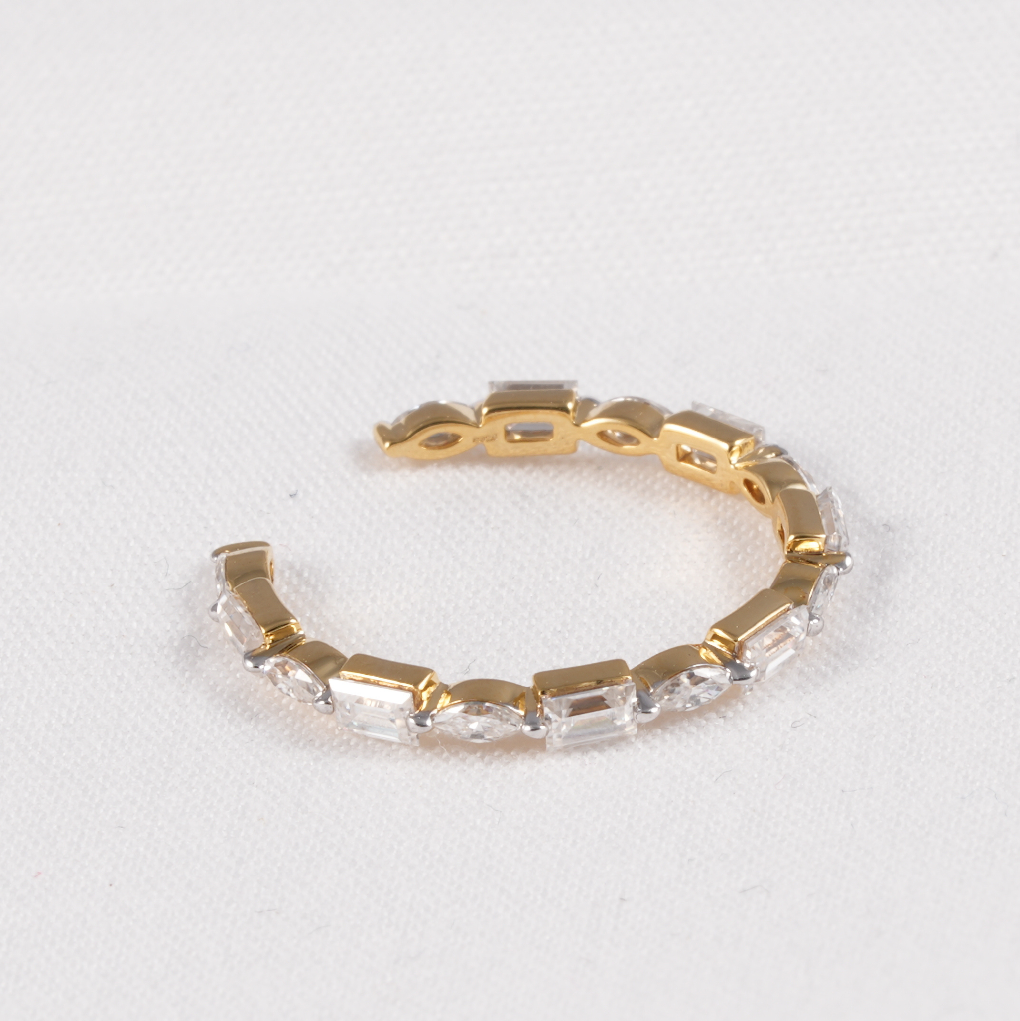 A gold-plated cuff bracelet featuring a row of clear baguette-cut crystals, displayed on a textured white background.