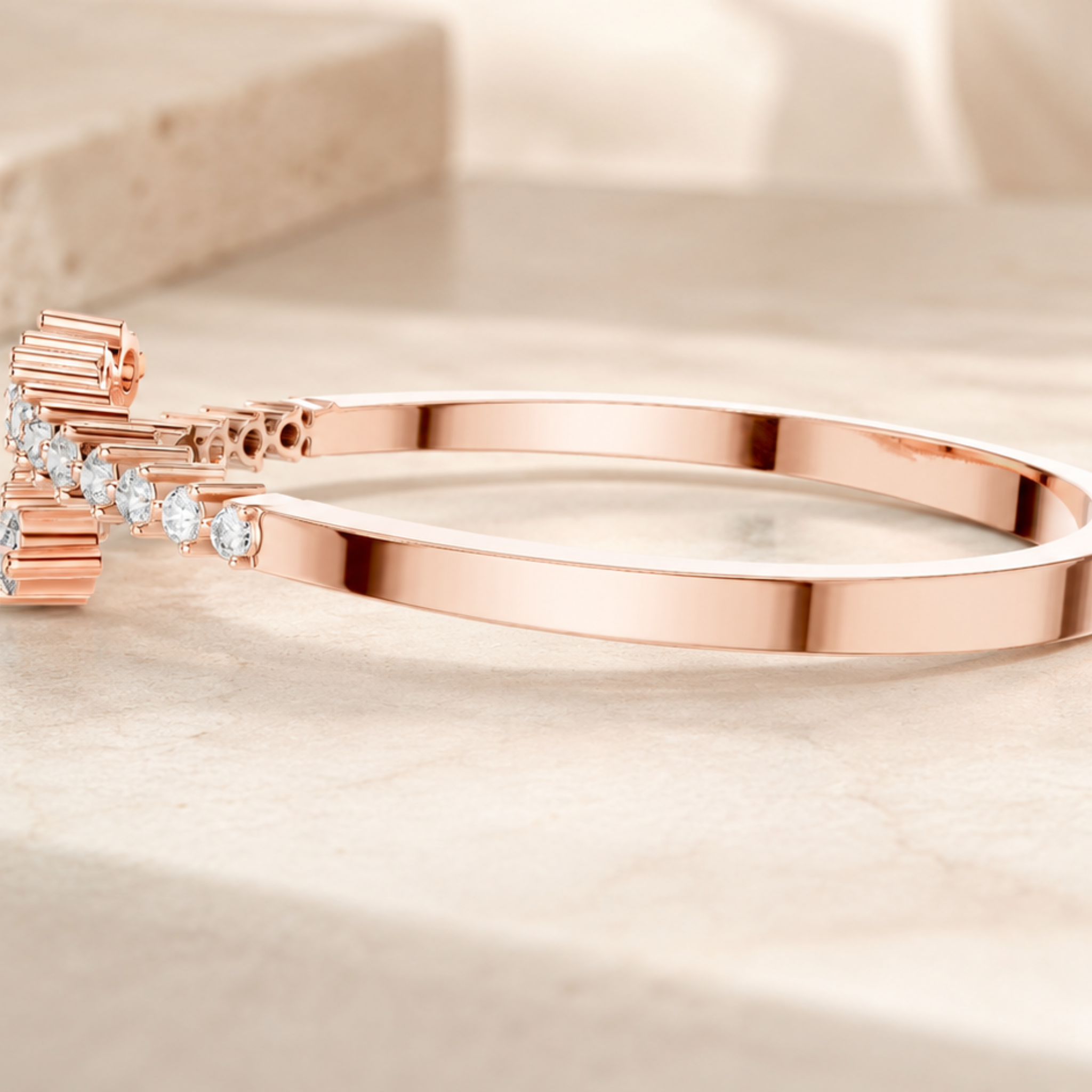 A polished rose gold bangle bracelet with a decorative clasp adorned with small clear stones, resting on a light beige marble surface.