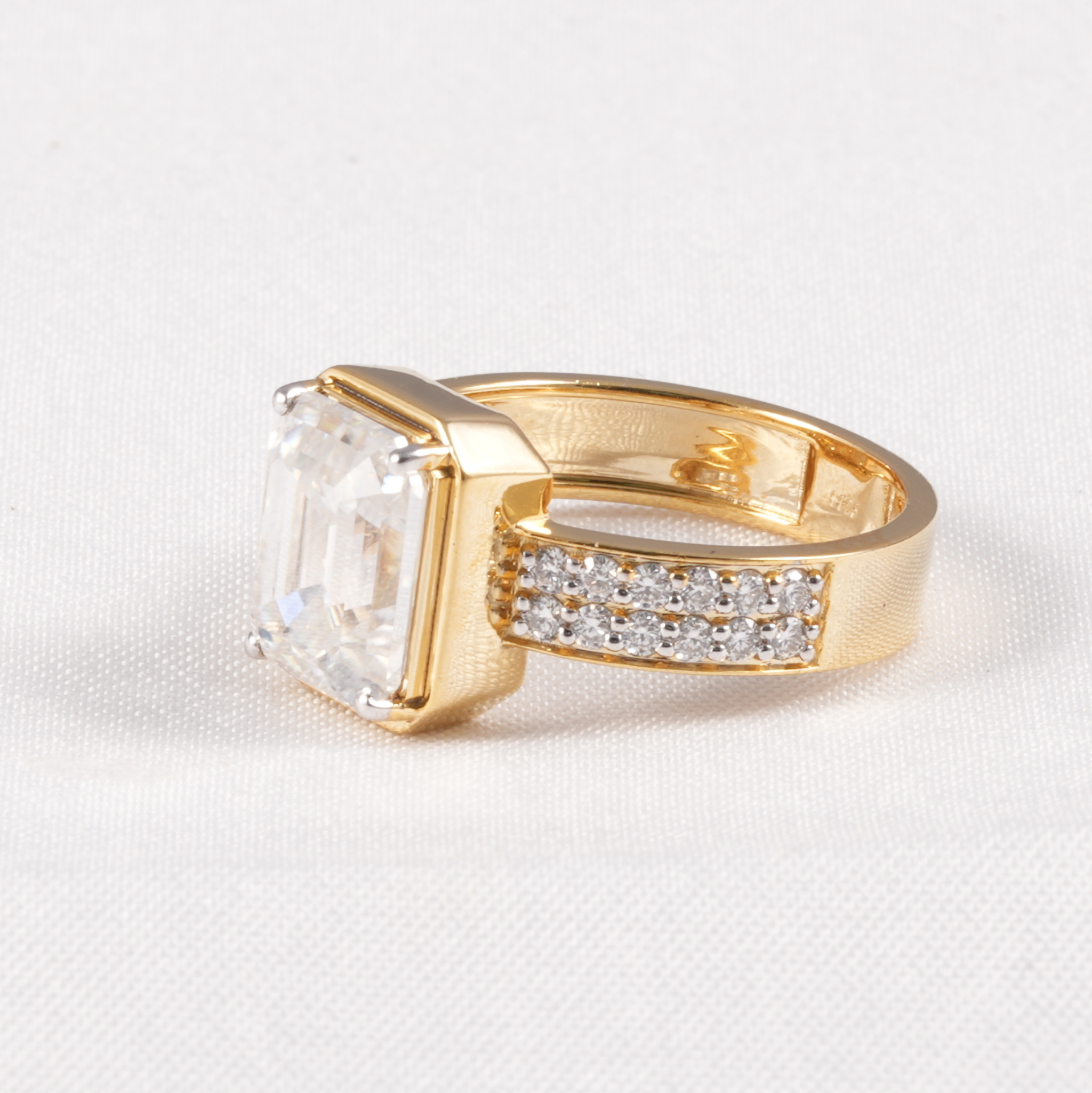 A gold ring featuring a large rectangular clear gemstone and a pavé band with small clear stones, photographed on a light textured fabric background.