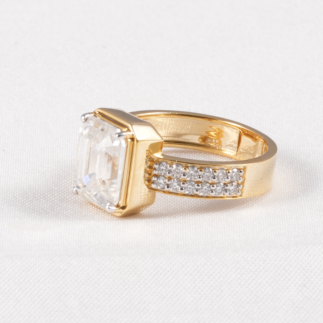 A gold ring featuring a large rectangular clear gemstone and a pavé band with small clear stones, photographed on a light textured fabric background.
