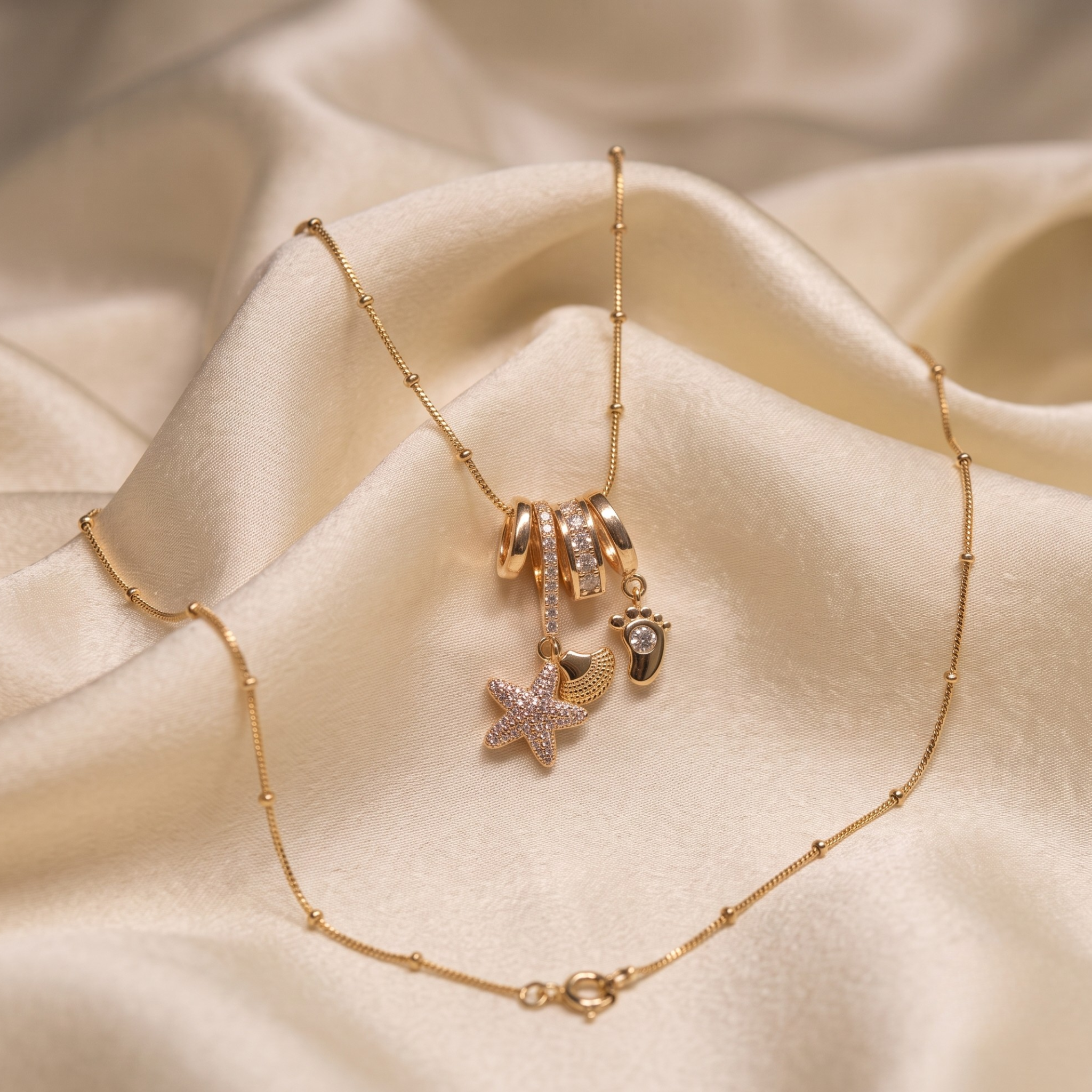 A gold-toned beaded chain necklace with charms, including a starfish, a small heart, and a footprint, displayed on draped cream satin fabric.