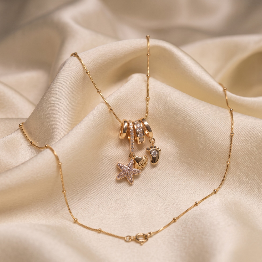 A gold-toned beaded chain necklace with charms, including a starfish, a small heart, and a footprint, displayed on draped cream satin fabric.