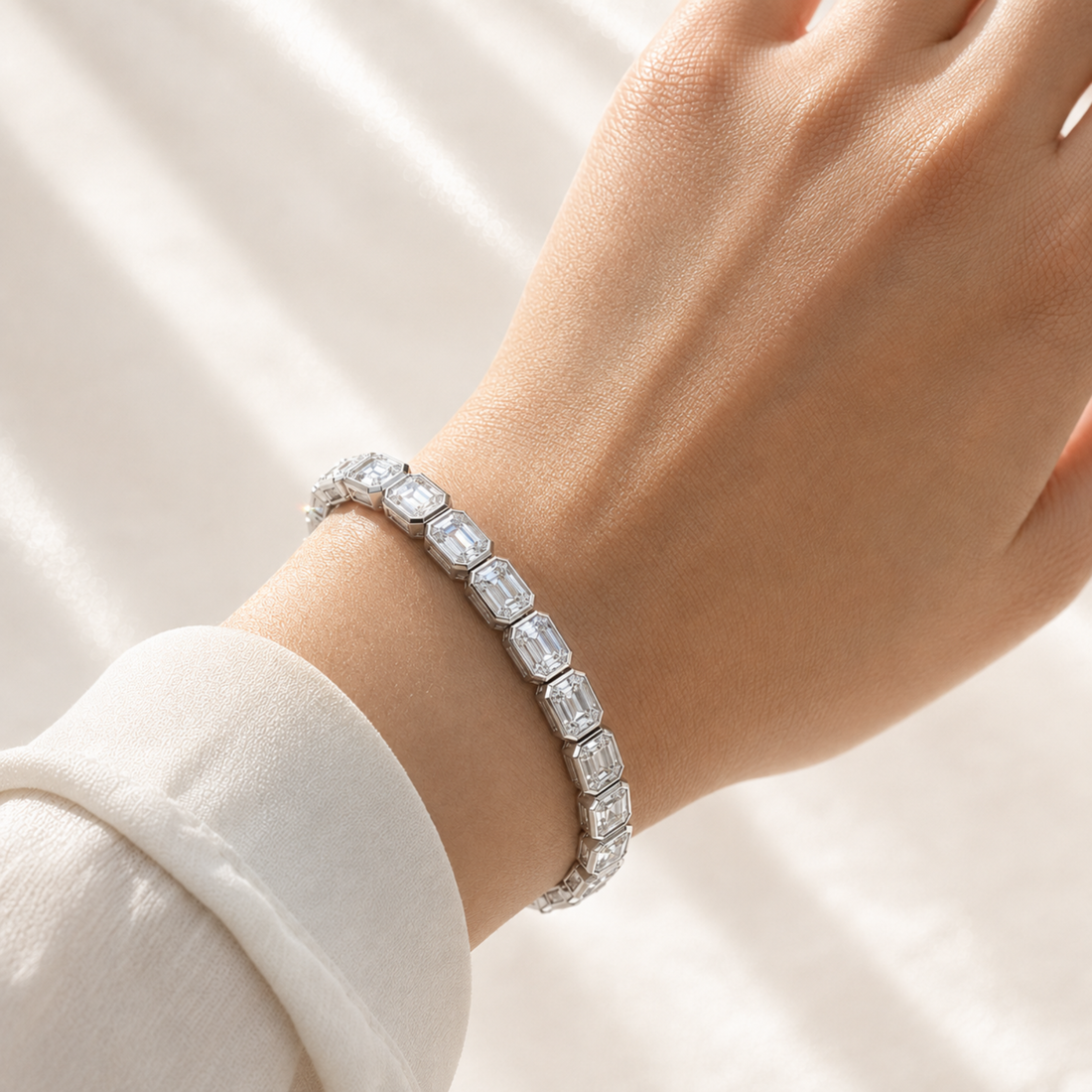 A close-up of a person's wrist wearing a silver tennis bracelet with Emerald-cut clear stones, set against a soft white draped fabric background.