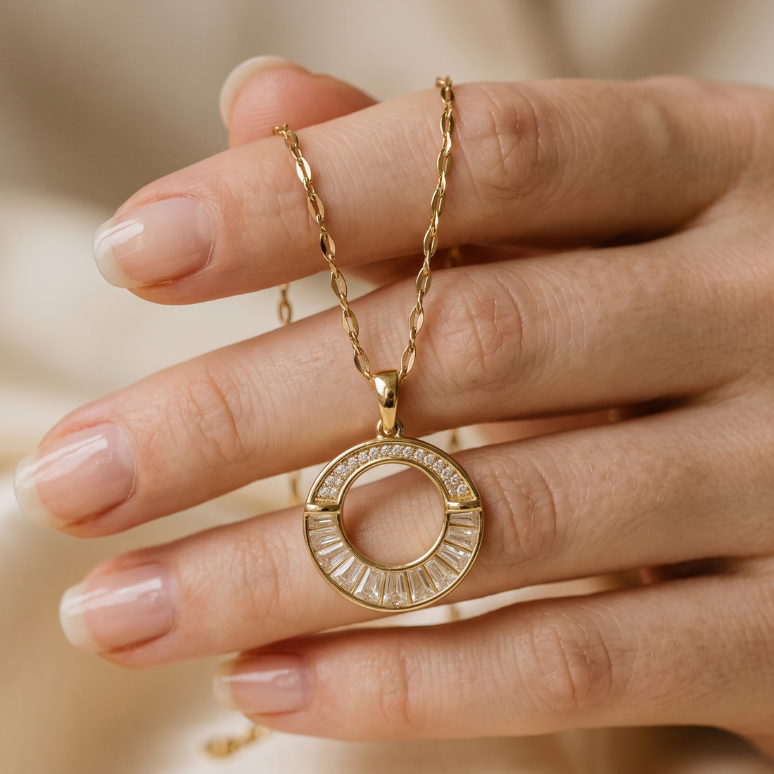 A gold necklace with a circular pendant featuring baguette-cut clear stones, held on a person's fingers against a soft beige background.