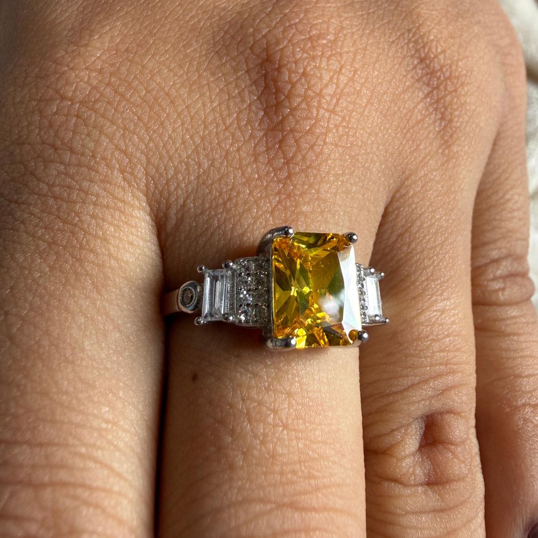 A close-up of a hand wearing a silver ring with a large yellow cushion-cut center stone and baguette side stones.