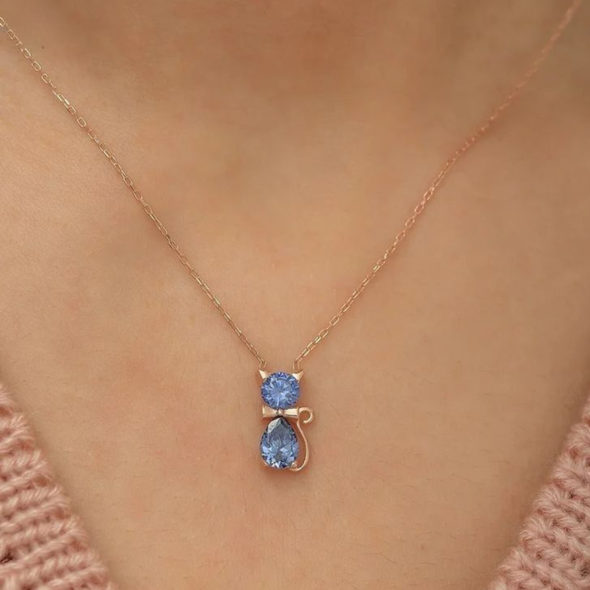 A close-up of a person's neck wearing a delicate rose gold necklace with a cat-shaped pendant featuring two light blue gemstones.