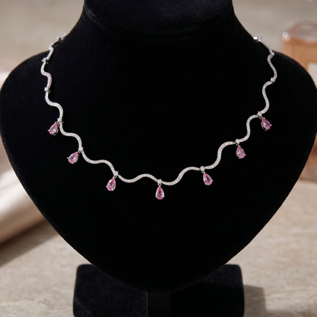 A wavy silver-toned necklace with small clear stones and dangling pink teardrop gemstones, displayed on a black velvet jewelry bust.