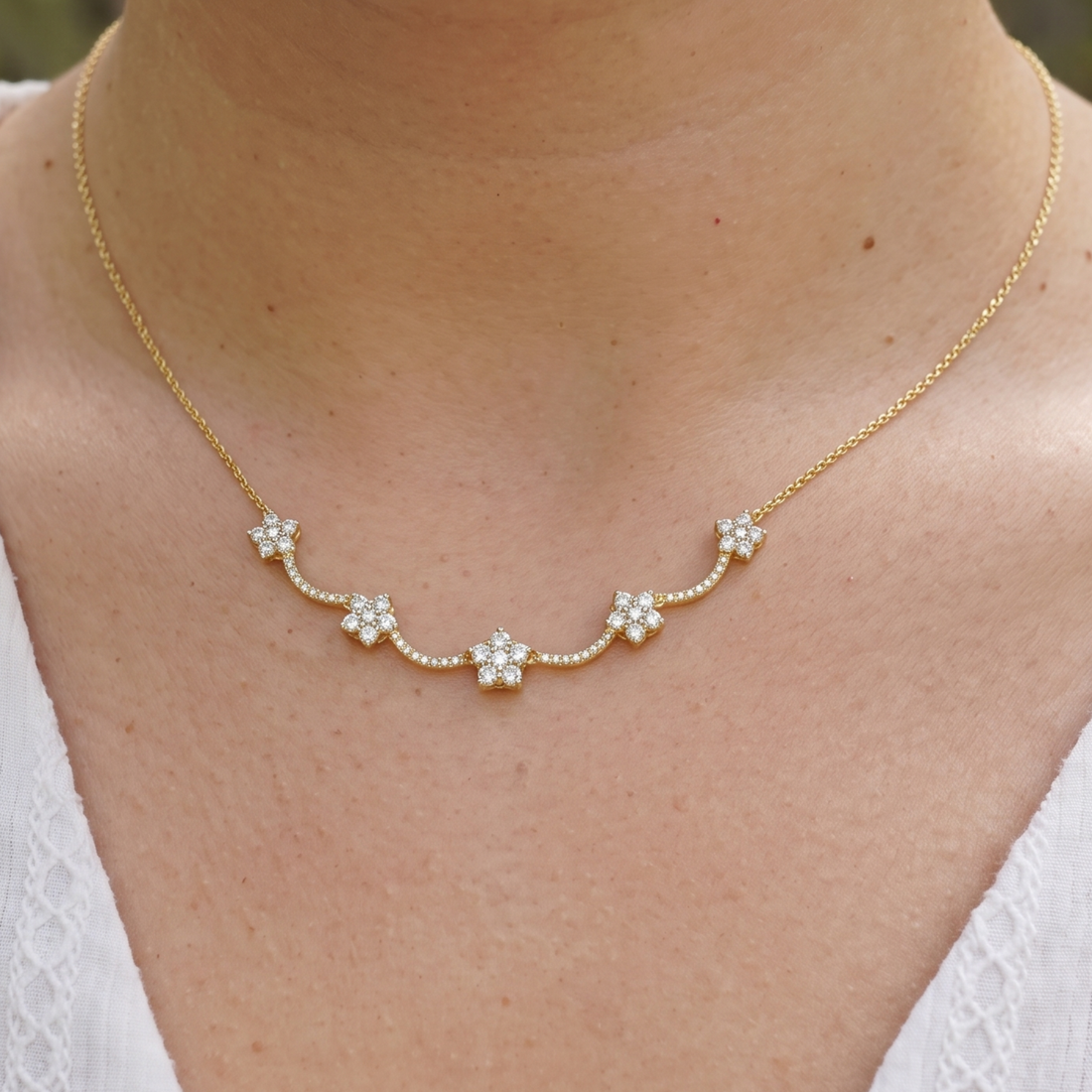 A delicate gold chain necklace with five small flower-shaped clusters of clear stones, worn on a model's neck over a white top.