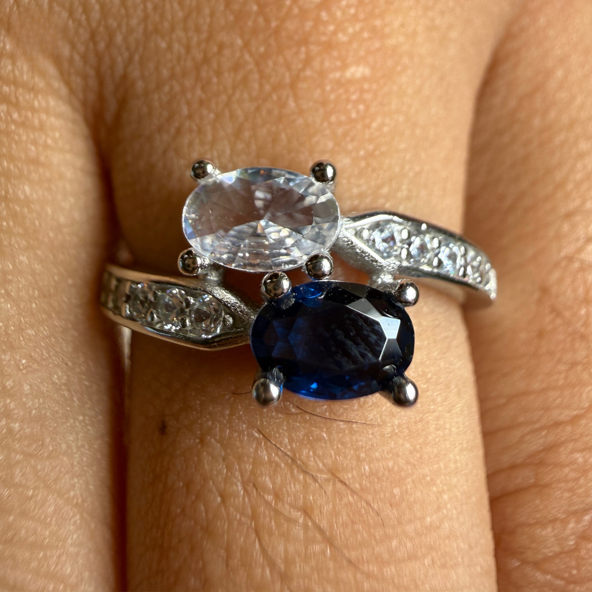 A silver ring with an oval clear gemstone and a larger oval deep blue gemstone, worn on a finger. The band is decorated with small clear stones.