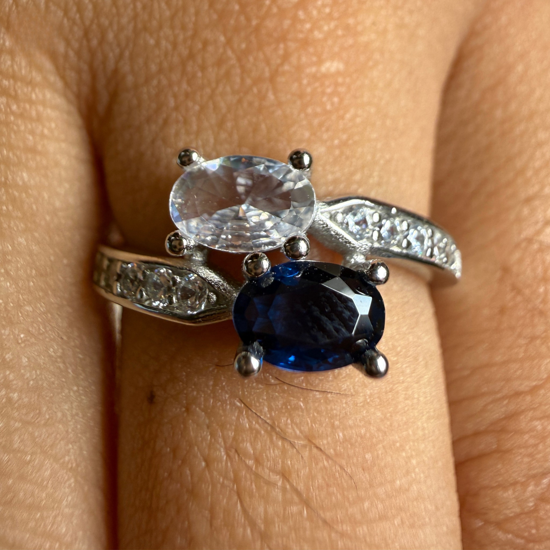A silver ring with an oval clear gemstone and a larger oval deep blue gemstone, worn on a finger. The band is decorated with small clear stones.
