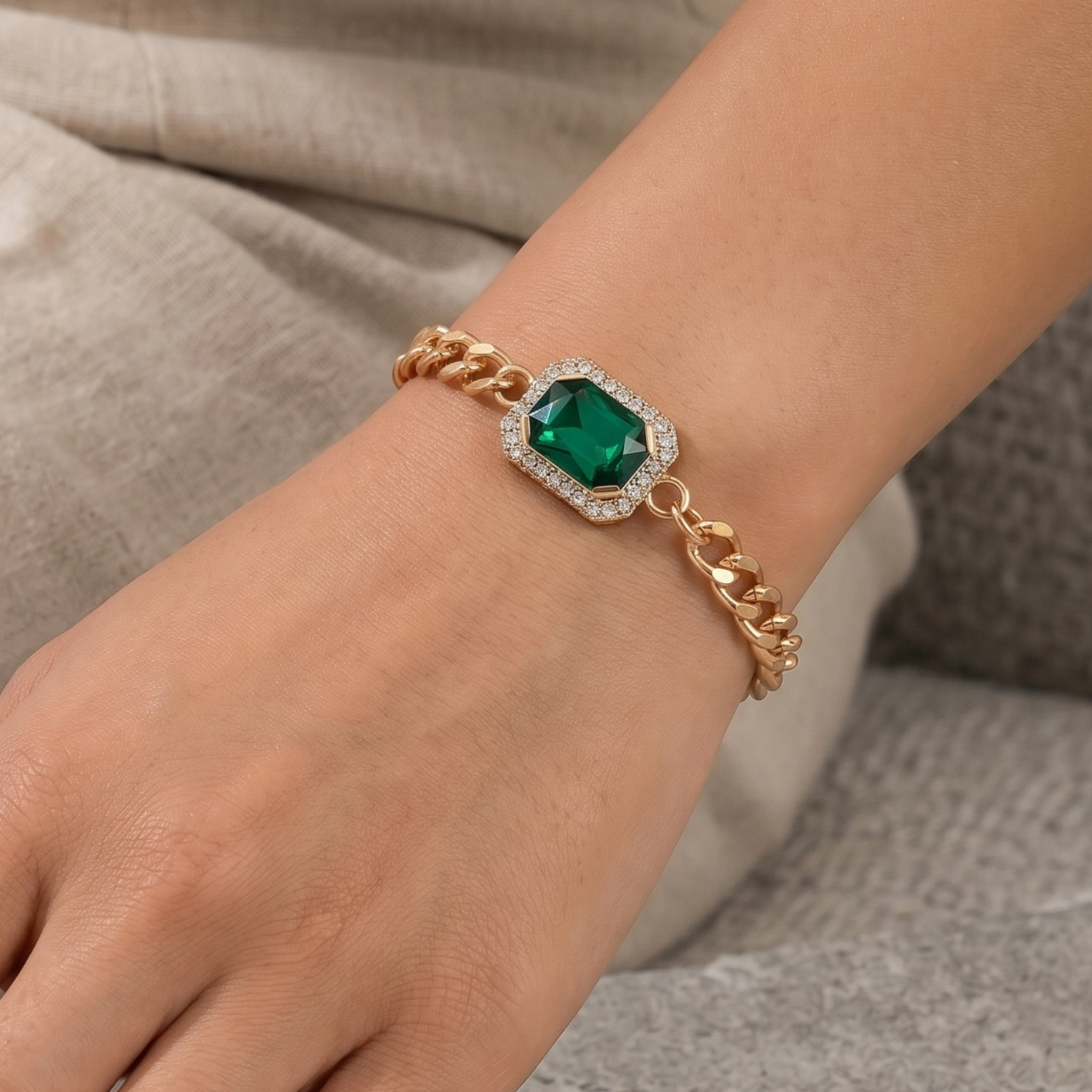 A close-up of a gold chain bracelet with a central green emerald-shaped gemstone surrounded by small clear stones, worn on a person's wrist.