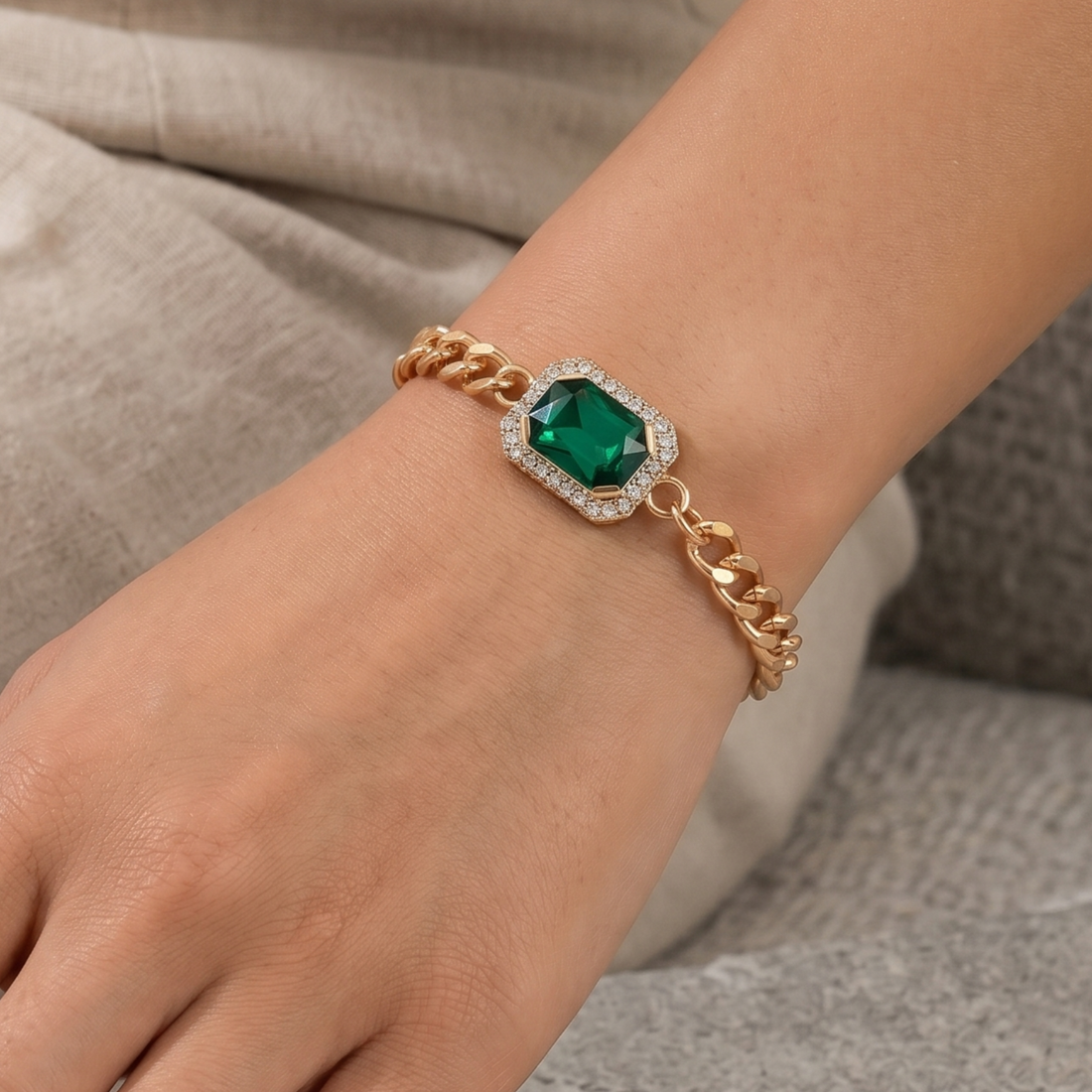 A close-up of a gold chain bracelet with a central green emerald-shaped gemstone surrounded by small clear stones, worn on a person's wrist.