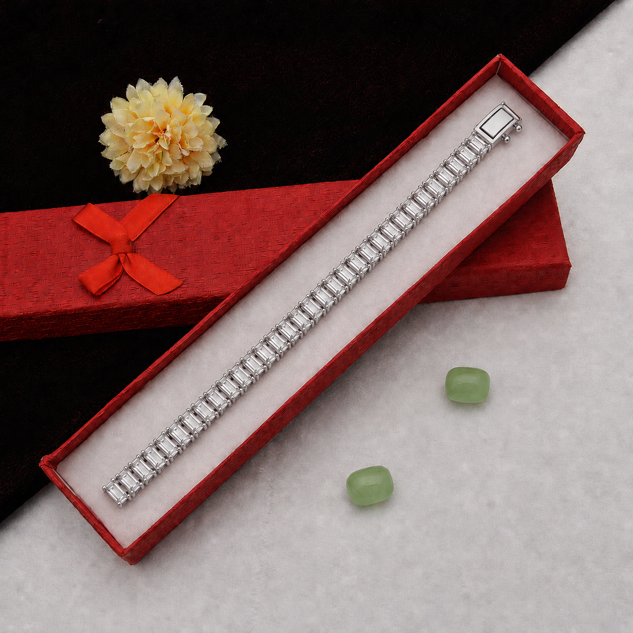 A silver-toned rectangular-link bracelet displayed diagonally inside an open red jewelry box, with a matching red box and ribbon in the background and two small green stones on a light surface.
