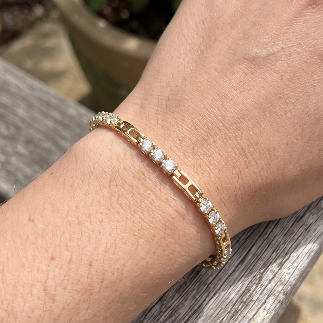 A gold tennis bracelet with clear round-cut gemstones worn on a person's wrist, resting on a weathered gray wooden surface.