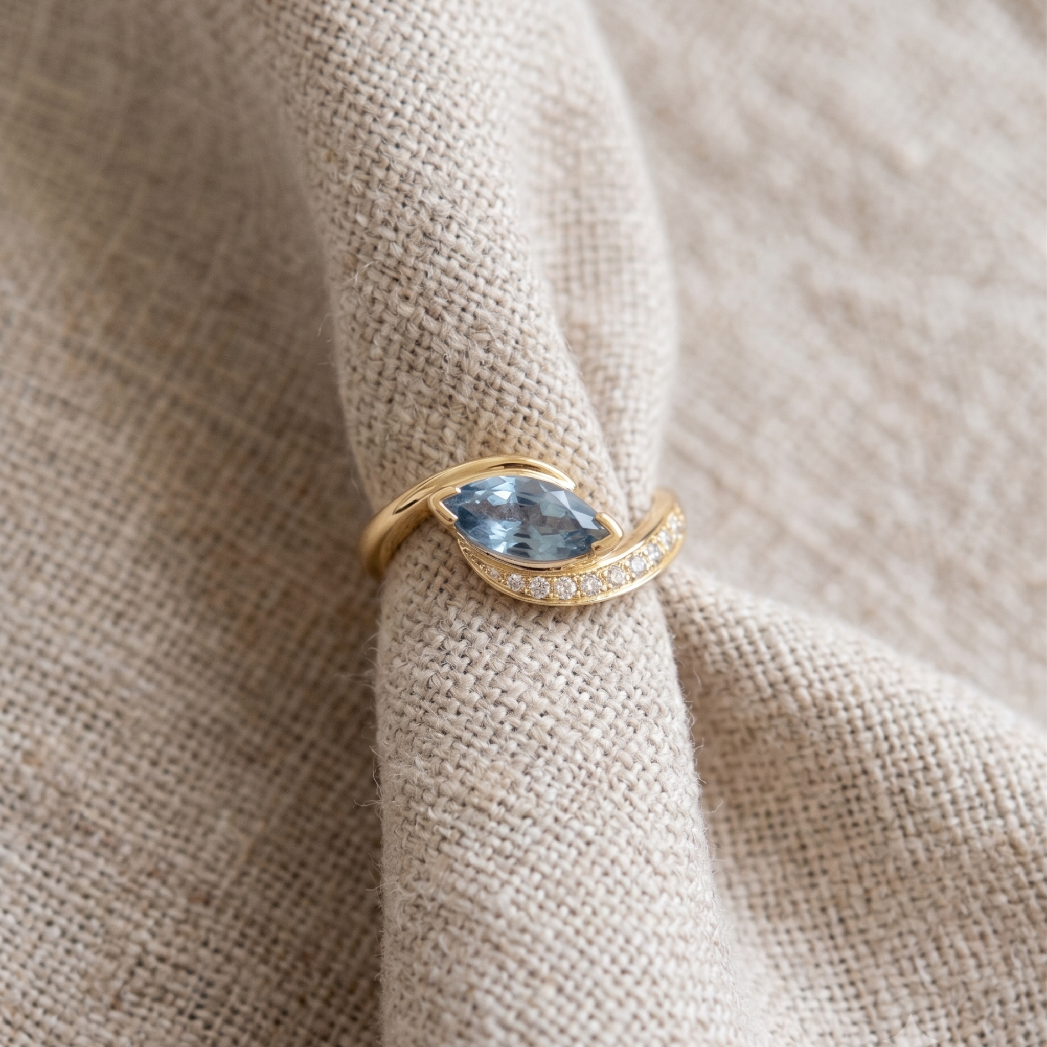 A gold ring with a marquise-cut light blue gemstone and a row of small clear stones, displayed on a textured beige fabric background.