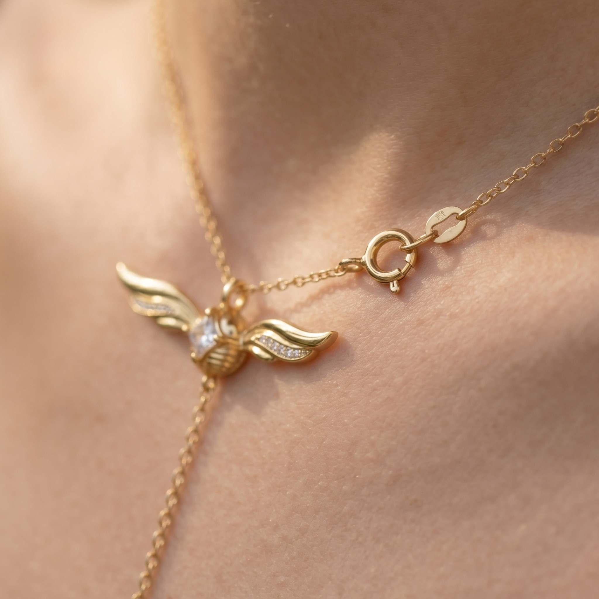 A close-up of a delicate gold chain necklace worn on skin, featuring a winged pendant with a small clear central stone and a lobster clasp.