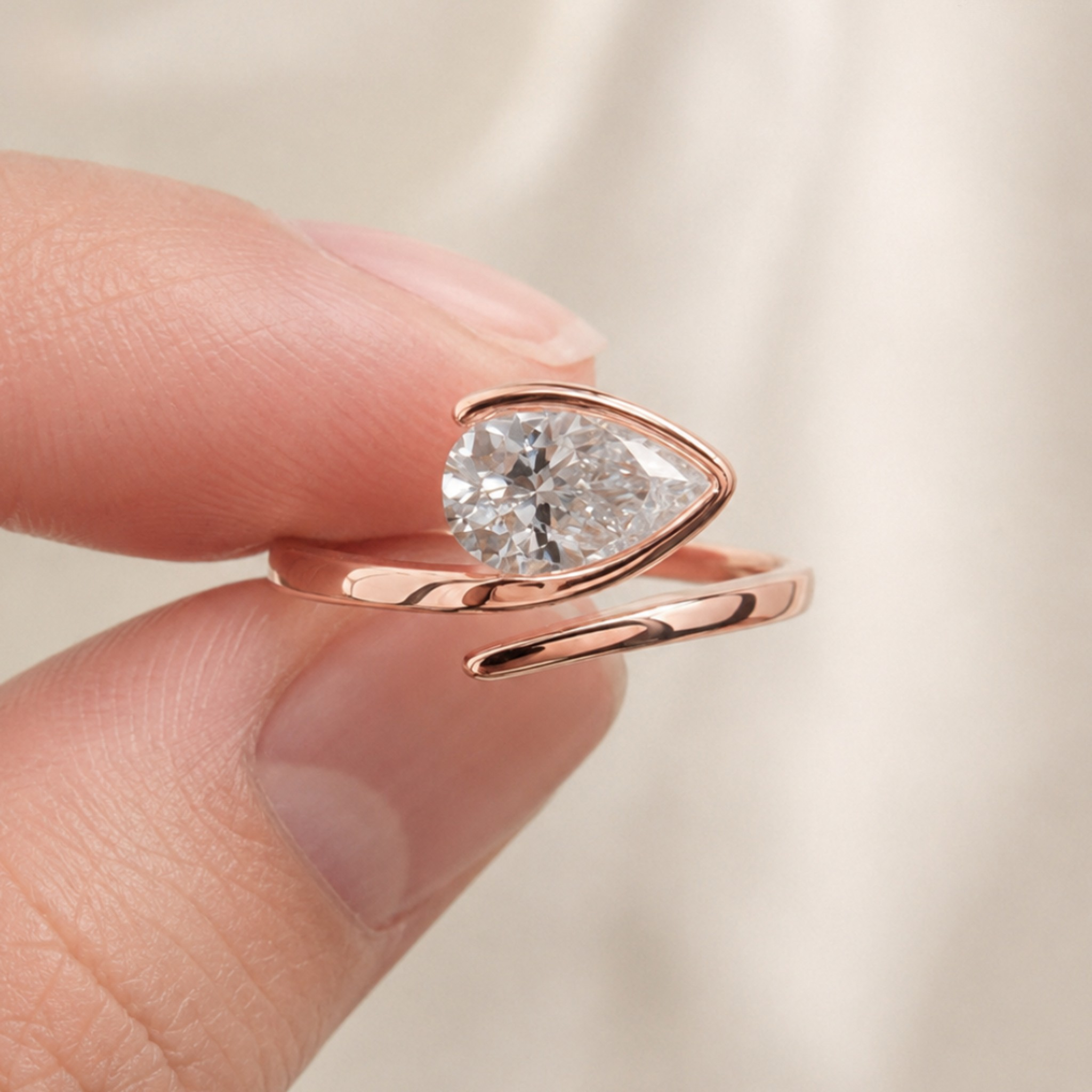 A close-up of a rose gold ring with a pear-cut clear gemstone, held between a person's fingers against a soft cream-colored fabric background.