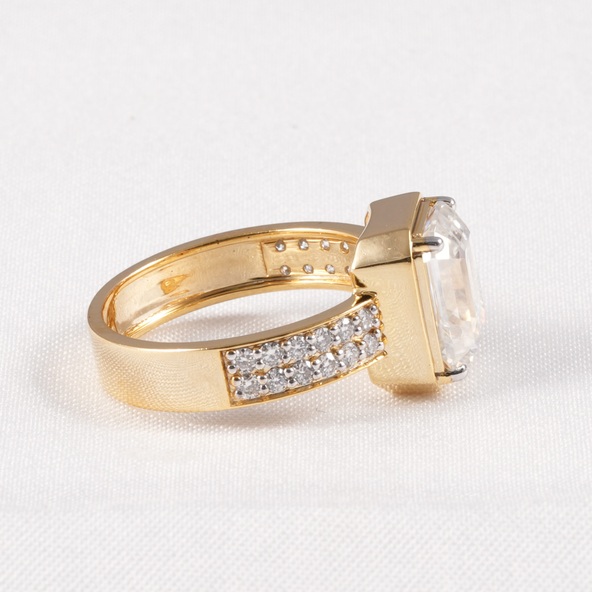 A yellow gold ring featuring a large rectangular clear gemstone and a pavé band set with small clear stones, photographed on a textured white fabric background.