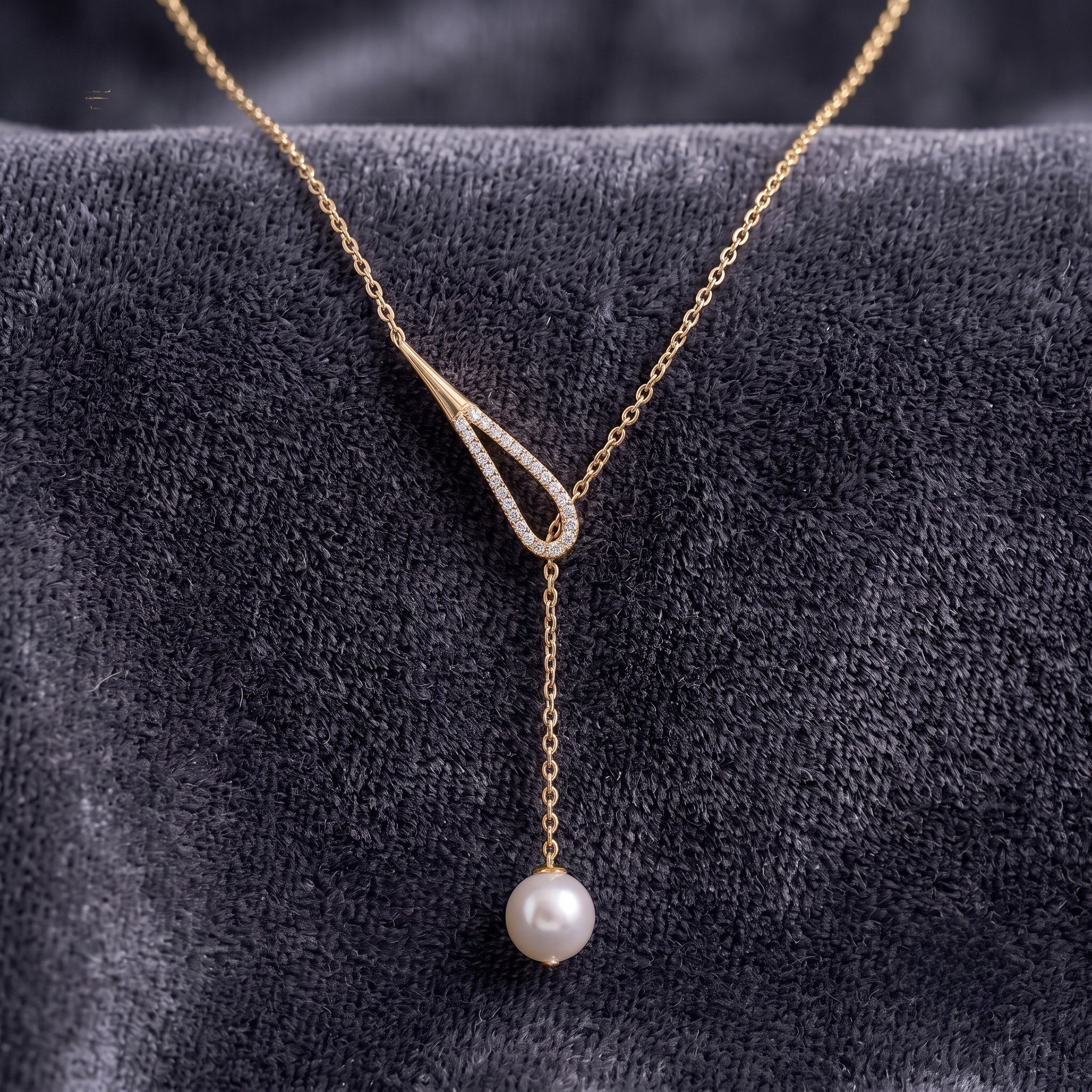 A gold Y-shaped necklace with a dangling white pearl, displayed on a textured dark gray velvet surface.