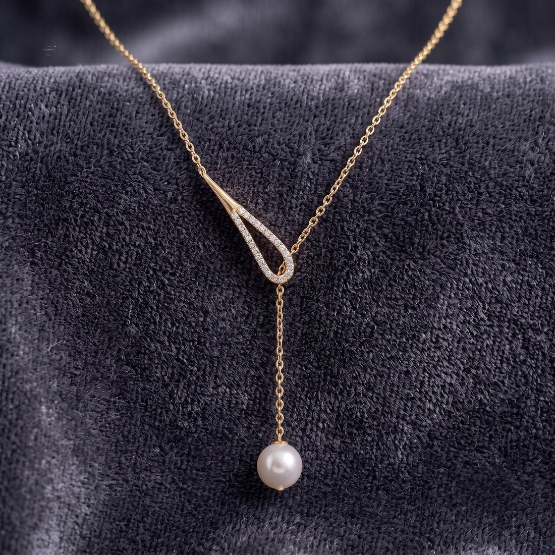 A gold Y-shaped necklace with a dangling white pearl, displayed on a textured dark gray velvet surface.
