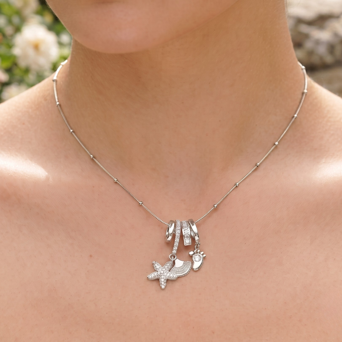 A silver charm necklace worn on a model's neck, featuring a starfish, footprint, and small animal silhouette charms on a delicate beaded chain.