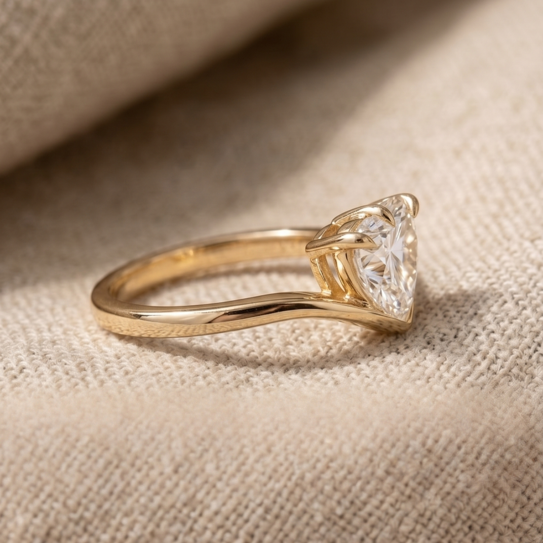 A gold solitaire ring with a triangular-cut clear gemstone, resting on a textured beige fabric background.