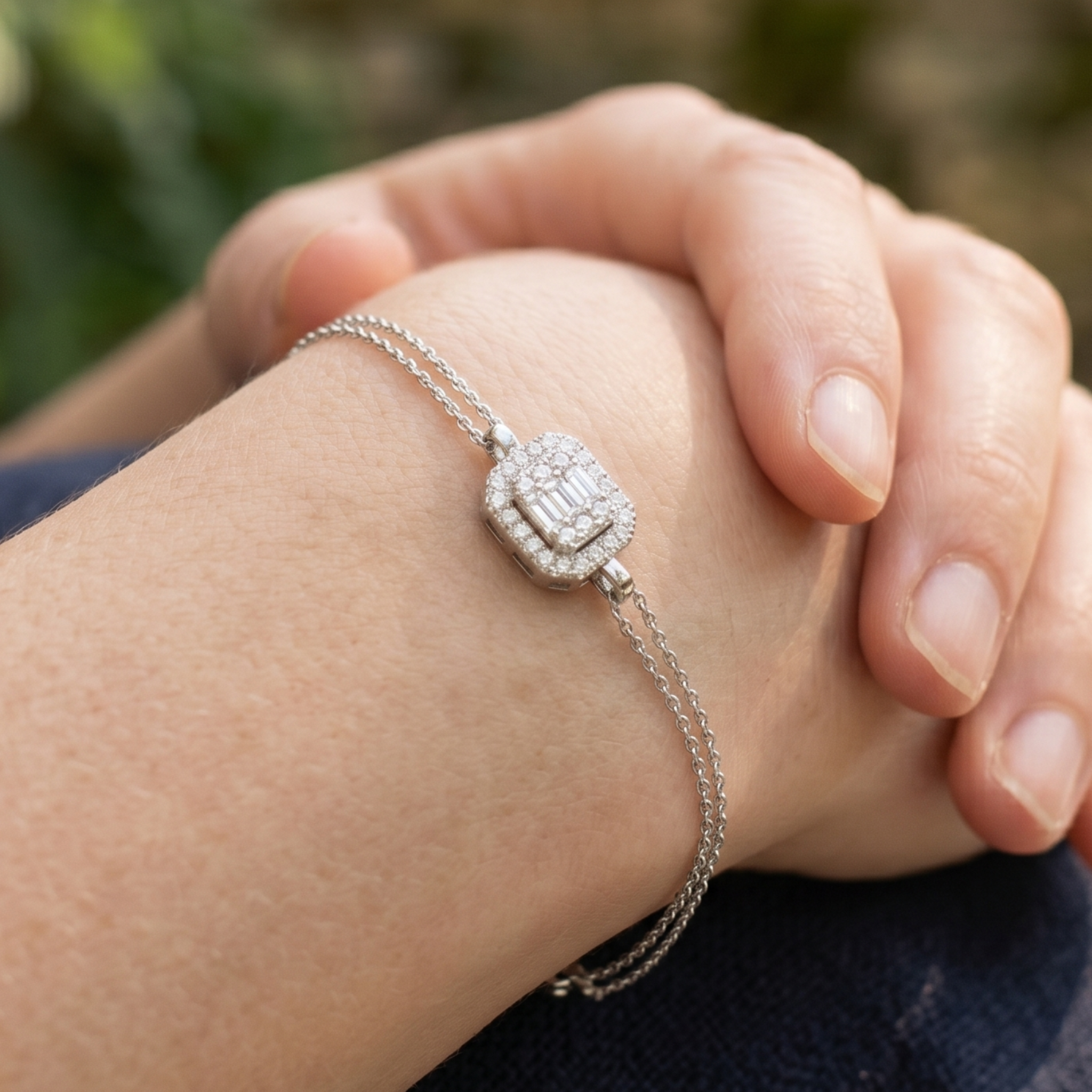 A close-up lifestyle shot of a delicate silver chain bracelet with a rectangular pavé diamond centerpiece, worn on a person's wrist with hands resting on their lap.
