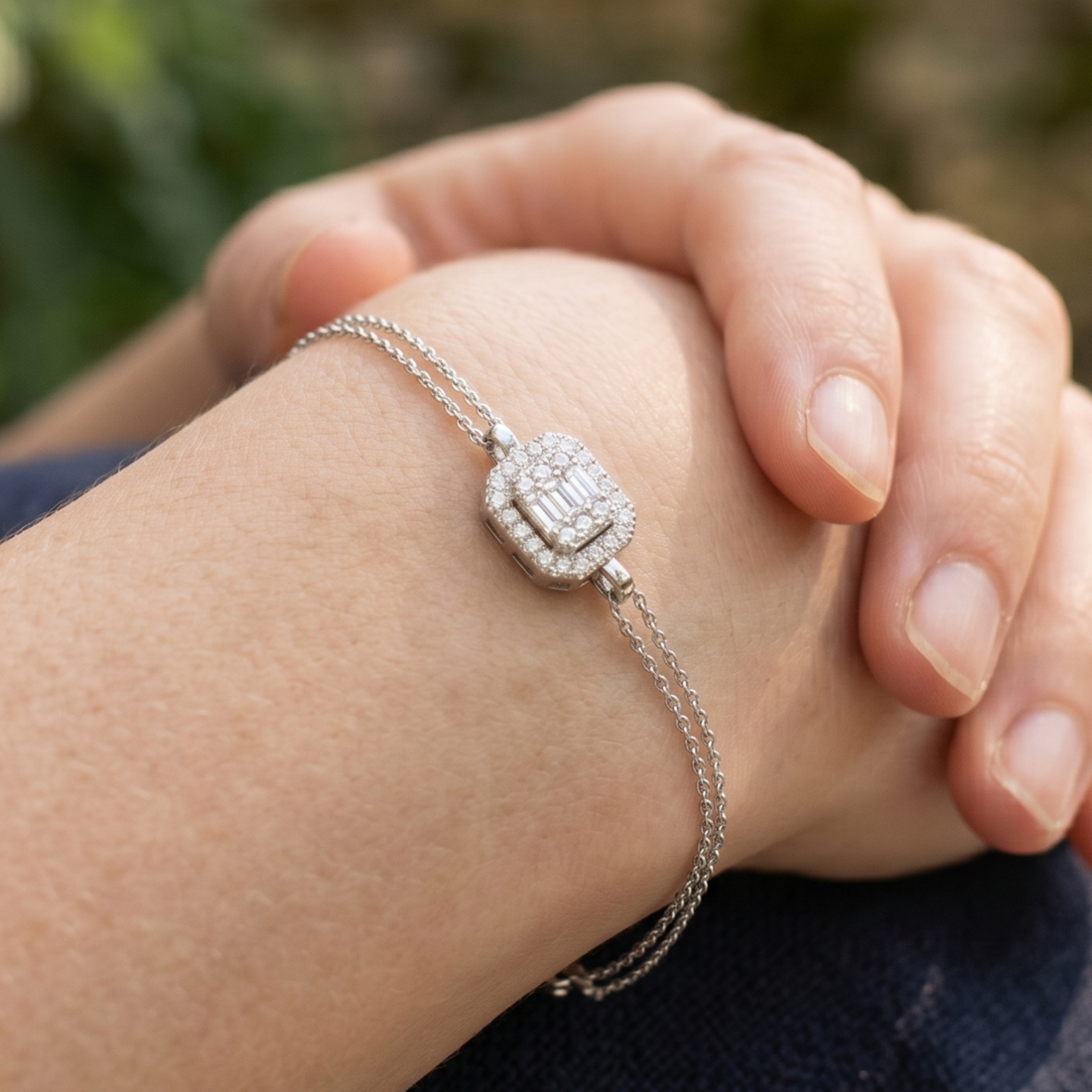 A close-up lifestyle shot of a delicate silver chain bracelet with a rectangular pavé diamond centerpiece, worn on a person's wrist with hands resting on their lap.