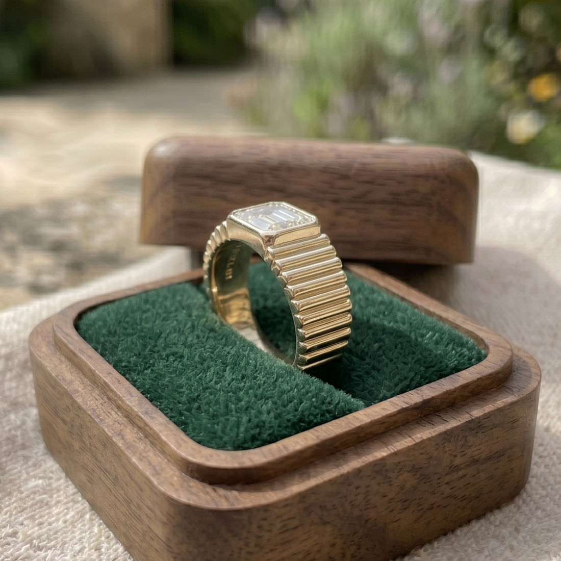 A gold ring with a square clear gemstone and a fluted band, displayed inside an open wooden box with a green velvet lining.