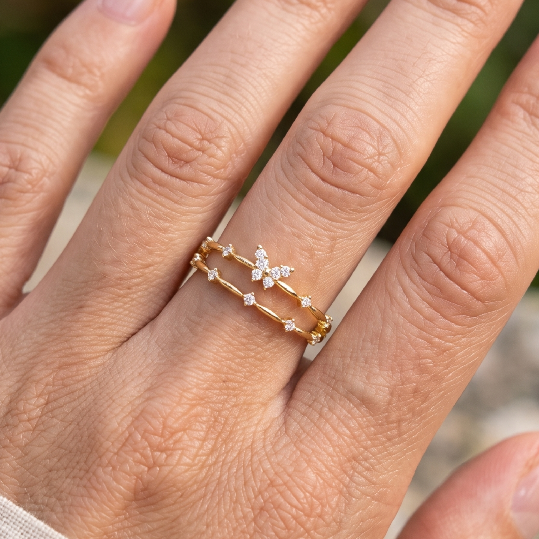 A close-up of a hand wearing two delicate gold rings. One ring features a small white floral cluster set with tiny clear stones, while the other is a thin gold chain with small star-shaped details.
