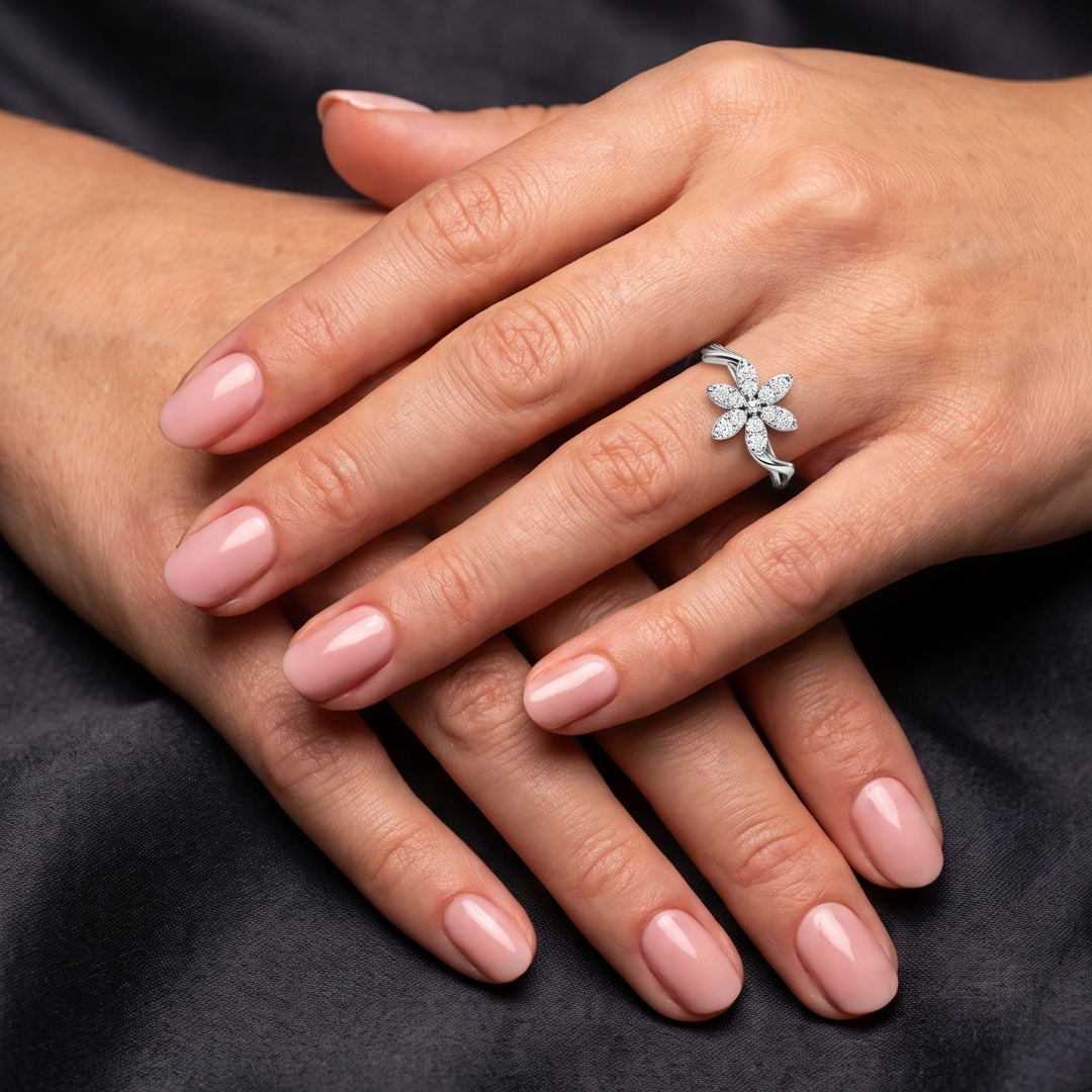 A close-up of crossed hands with a pale pink manicure, wearing a silver flower-shaped ring set with clear stones, resting on a dark fabric background.