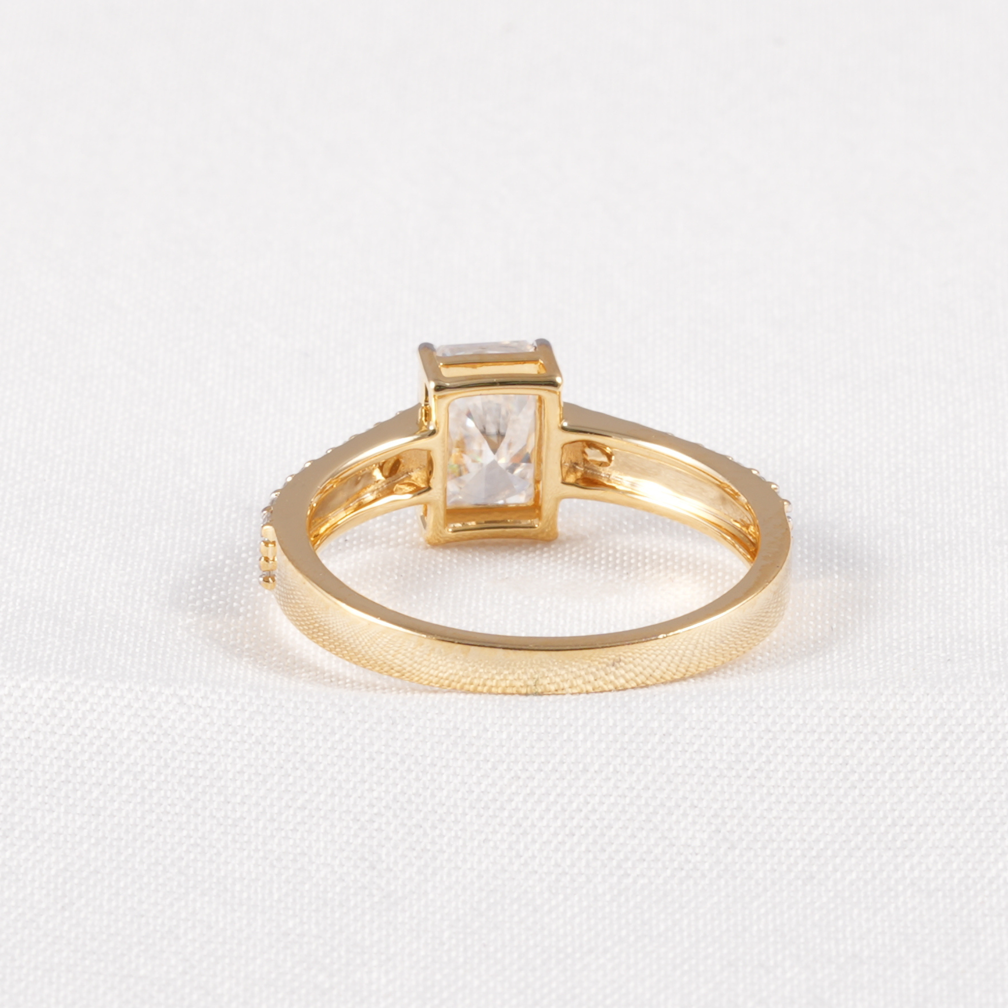 A gold ring with a rectangular clear gemstone in a bezel setting, displayed on a textured white fabric background.