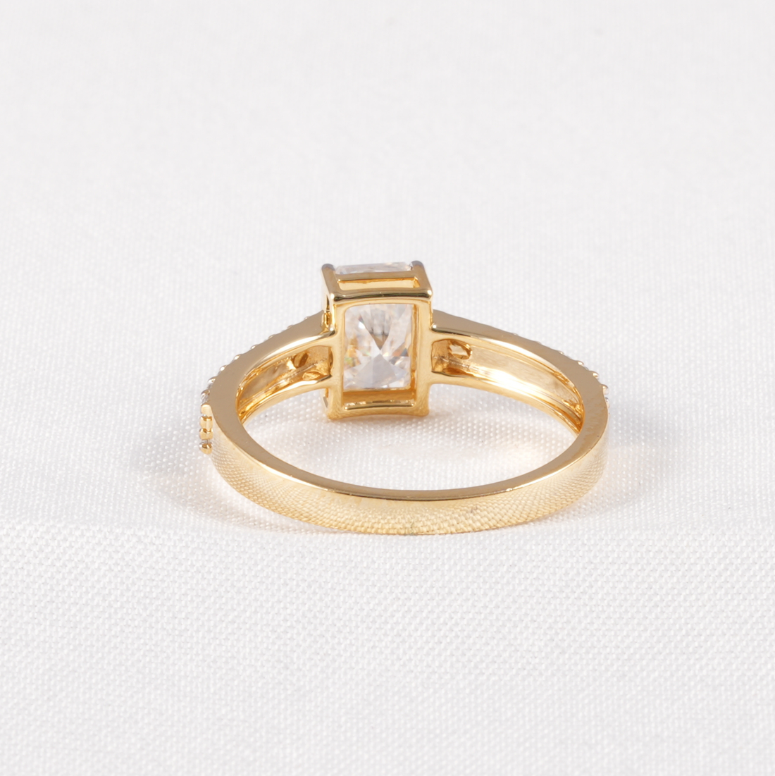 A gold ring with a rectangular clear gemstone in a bezel setting, displayed on a textured white fabric background.