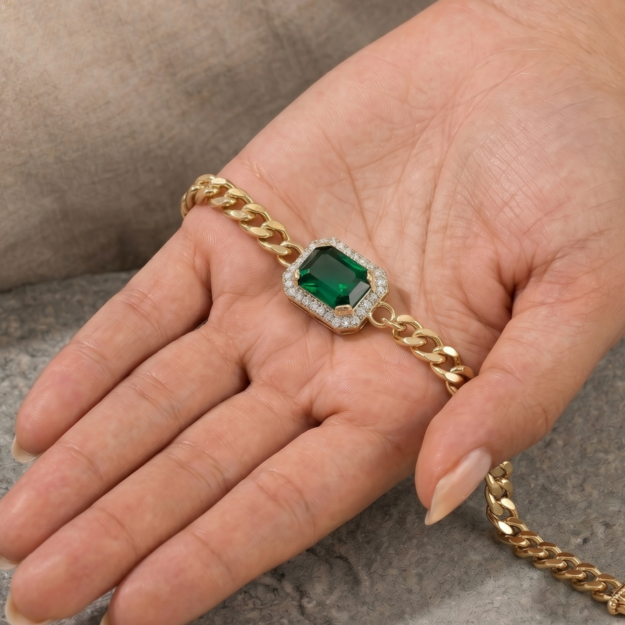 A gold chain bracelet with a central emerald-cut green gemstone surrounded by a halo of small clear stones, displayed on a person's hand.